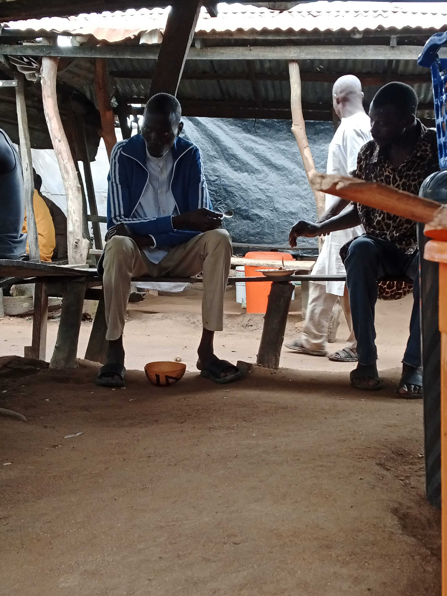 In the city of Unknown, Unknown, two men sit at a wooden table under a rustic, open-air shelter with a corrugated metal roof supported by wooden posts. The man on the left, wearing a blue jacket with white stripes and light-colored pants, is seated and looking down. In front of him, on the ground, is an orange bowl with a dark design. The man on the right, dressed in a leopard-print shirt and dark pants, is also seated and appears to be interacting with something on the table. A third man, wearing a white t-shirt, stands in the background, facing away from the camera, and a fourth person is partially visible on the far left. The environment is earthy, with dirt flooring and a dark tarp visible behind the seated individuals. An orange plastic container and a plate with food are on the table.