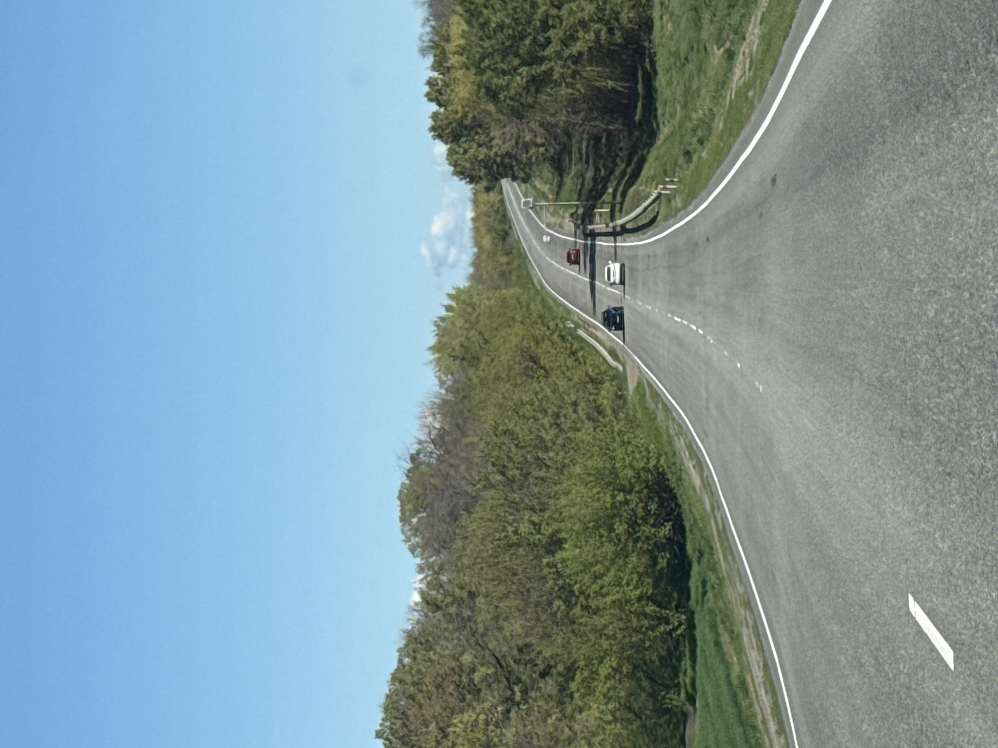 A view from above depicts a grey asphalt road curving through a rural landscape under a clear blue sky. Lush green trees and grass line the sides of the road. Several vehicles are seen driving on the road. The location appears to be Pokrovskoye, Russia, based on details like the type of foliage and the style of road.