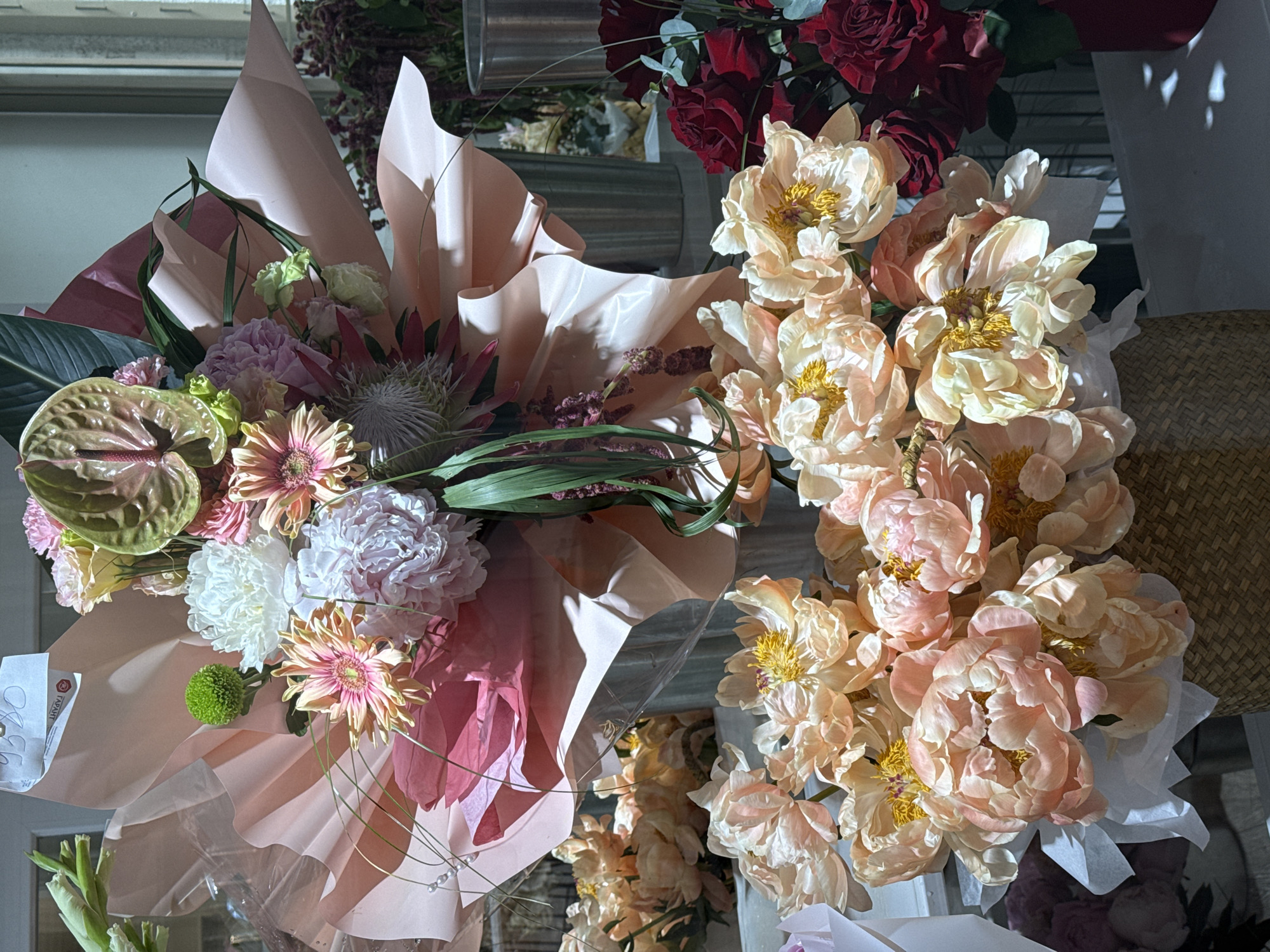 This close-up image captures a vibrant display of flower bouquets, likely within a florist shop or market setting. The scene is dominated by two distinct floral arrangements.
On the left, a large, elaborate bouquet is wrapped in layered pink and darker, almost reddish, paper. This bouquet features a diverse assortment of flowers, including a prominent green and pink anthurium, a spiky protea, several pink gerbera daisies, a few white and light pink peonies, and a small, spherical bright green flower that resembles a button mum or similar bloom. Various green leaves and delicate foliage are interspersed throughout the arrangement. A white tag with partially visible text, including "ГАРАНТ" and "OBO," is tucked into the wrapping.
To the right, another substantial bouquet is almost entirely composed of numerous light peach and pale pink peonies, all with distinct yellow centers. This arrangement is wrapped in white paper, providing a contrast to the more colorful left bouquet.
In the background, hints of other floral displays are visible, including a cluster of dark red roses in the upper right quadrant. A light brown wicker-like texture, possibly a basket or stand, can be seen on the far right. Metallic cylindrical objects, likely vases or containers, are also present in the blurred background, reinforcing the commercial floral setting.
The lighting is strong and directional, casting pronounced shadows on the wrapping paper and the petals of the flowers, which adds depth and texture to the image. The flowers appear fresh and in full bloom, suggesting a lively and well-maintained display. The overall impression is one of beauty, abundance, and intricate floral artistry.