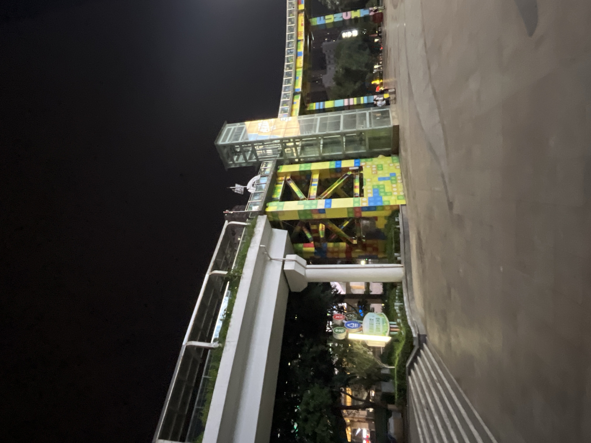 This image captures an outdoor scene at night in Jiulongpo District, China, near a multi-level pedestrian walkway decorated with colorful, block-like patterns. The walkway, elevated above the ground, appears to be part of an urban entertainment or recreational area. A glass elevator structure is visible, integrated into one of the support columns for the walkway. To the left, a sign with various icons and text in Chinese is illuminated. The ground in the foreground is paved, with a set of stairs leading up to the walkway on the left side. Trees and some buildings are visible in the background, illuminated by artificial lights. A few people are present, walking in the distance. The overall impression is of a lively urban space at night.