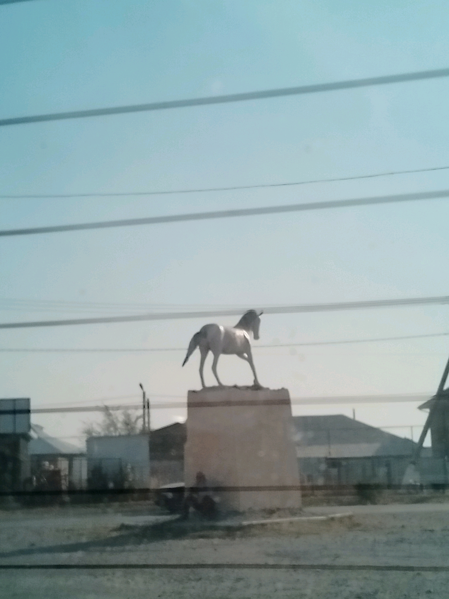 A white horse statue stands on a beige pedestal in Көлкент, Kazakhstan. The statue faces to the left, with its head lowered and its tail raised. The horse is in a static, posed position, suggesting it is a monument. The pedestal is rectangular and appears weathered. In the background, there are several buildings, some of which have dark roofs. There are also power lines visible, crisscrossing the frame horizontally. The sky is a pale blue, with some horizontal lines that may be reflections on a window. The overall scene appears to be captured through a window, as evidenced by the reflections and the slightly blurry quality of the image. The ground in front of the pedestal is a sandy or gravelly area. The lighting suggests it is daytime, possibly late afternoon or early morning, given the soft light.