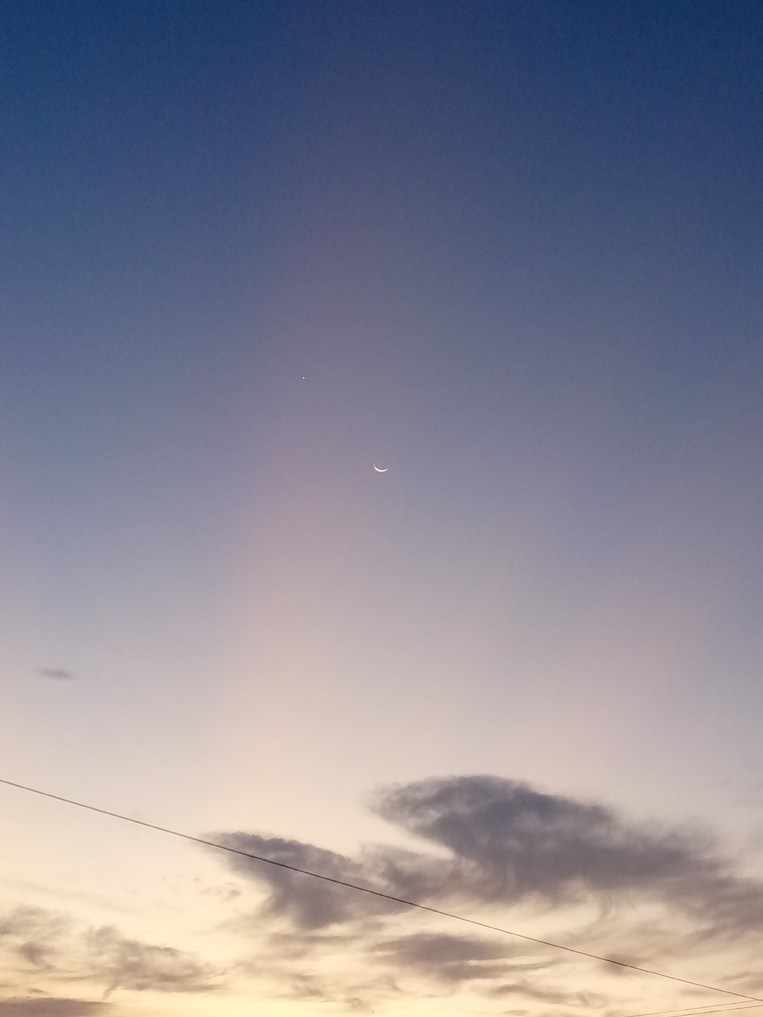 A twilight sky over Las Guevaras, Venezuela. A crescent moon is centered in the upper middle portion of the frame. Above and slightly left of the moon is a single bright star or planet. Below the moon and scattered near the bottom of the frame are dark clouds. A power line runs diagonally across the lower portion of the sky. The sky transitions from a dark blue at the top to a lighter peach/orange at the horizon.