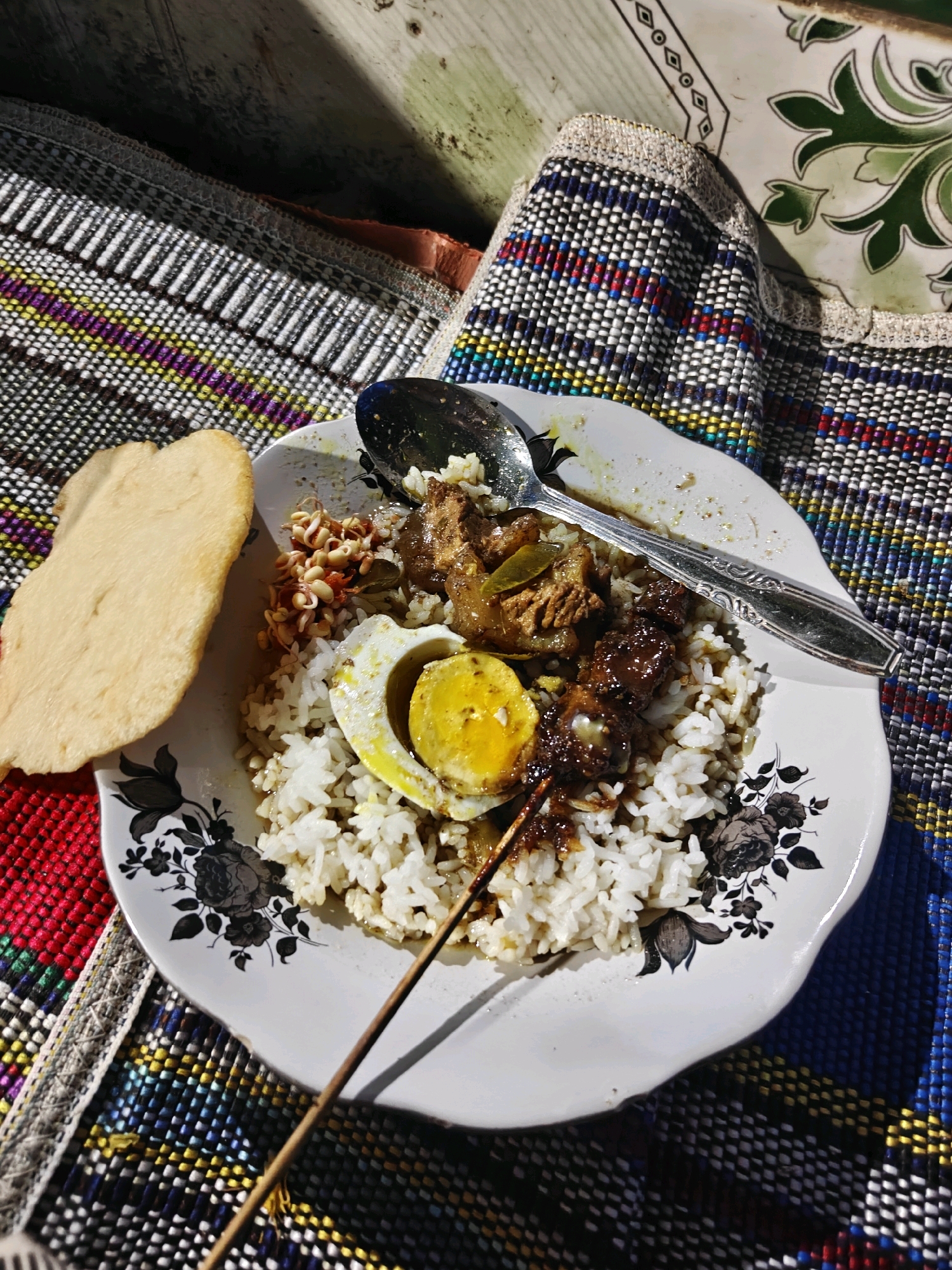 A top-down shot shows a plate of food on a woven mat. The plate is white with black floral designs on the rim and contains white rice, a hard-boiled egg cut in half with the yolk facing up, some meat in a dark sauce, and a small pile of sprouts. A spoon rests on the plate, and a wooden skewer is stuck into the rice and meat. To the left of the plate is a single, light-colored, irregular-shaped cracker. The background consists of a wooden surface and the colorful woven mat. The lighting suggests it is daytime, with shadows cast across the scene, indicating natural light. The image does not provide any specific indicators of location within Cermee, Indonesia.
