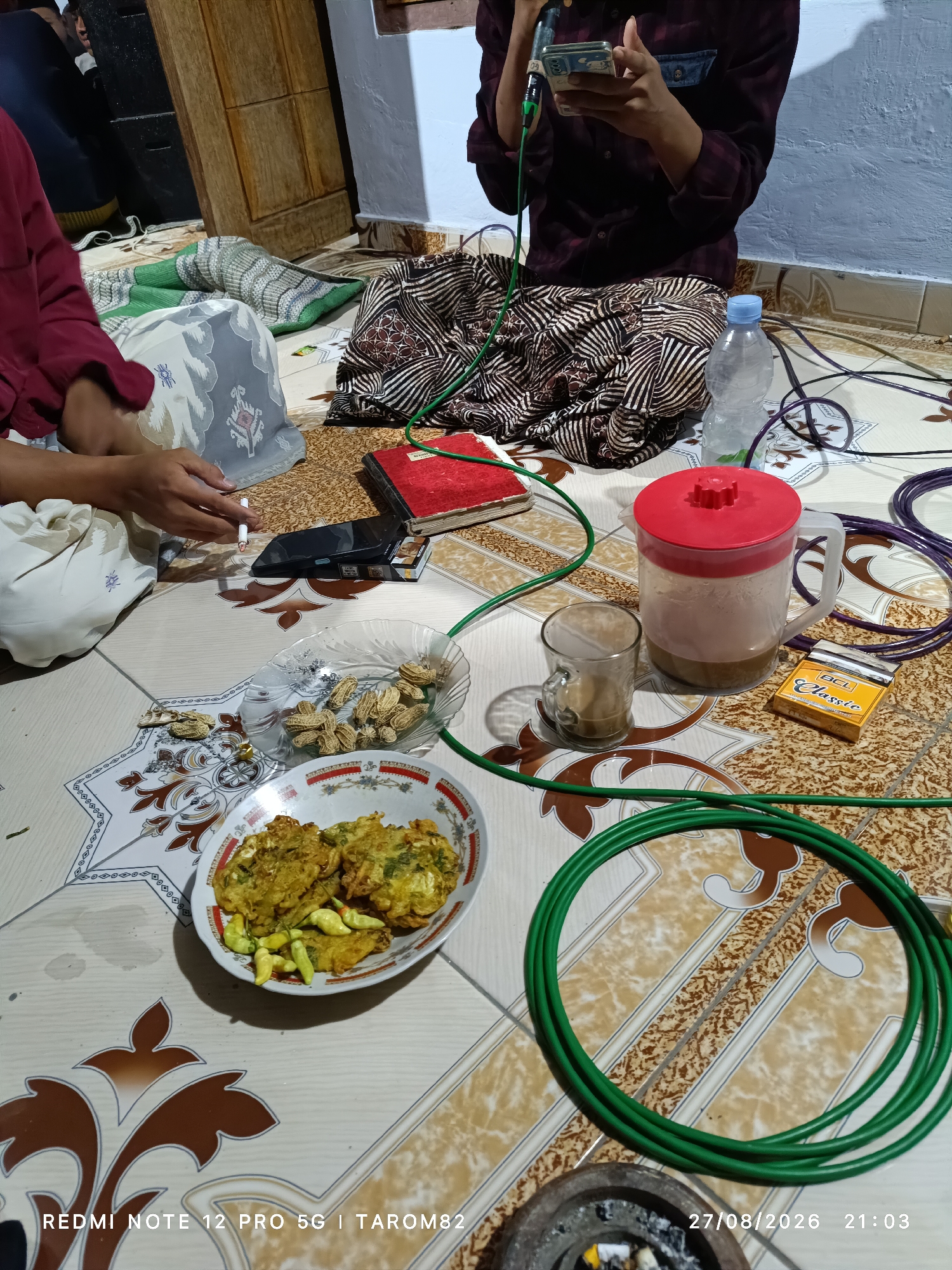 The image captures a casual indoor scene, likely a home gathering, in Rejoagung, Indonesia. The focus is on a collection of objects spread across a patterned tiled floor, suggesting people are sitting or lying on the floor.
In the foreground, a white bowl contains several pieces of fried food, possibly fritters or tempura, accompanied by a small pile of light-colored chilies. To the left of this bowl is a smaller glass plate filled with peanuts in their shells. A person's hands are visible in the lower left corner, one holding a lit cigarette and the other appearing to be in motion. Next to the cigarette is a smartphone and a pack of "BCL Classic" cigarettes.
Further back, a clear plastic water bottle stands upright. Beside it is a glass containing a brown beverage, likely coffee, and a large pitcher with a red lid also contains a similar brown liquid. A red-covered book lies open on the floor. In the upper right portion of the image, a person wearing a dark patterned shirt and skirt is seated, holding a smartphone and what appears to be a microphone or recording device.
The floor is covered in light-colored tiles with a repeating brown and tan pattern. Green and purple cables are coiled on the floor, suggesting the presence of electronic equipment. The overall lighting appears to be artificial, casting shadows. The timestamp in the lower right corner indicates "27/08/2026 21:03". The presence of the people, food, drinks, and smoking materials suggests a social gathering or a break.