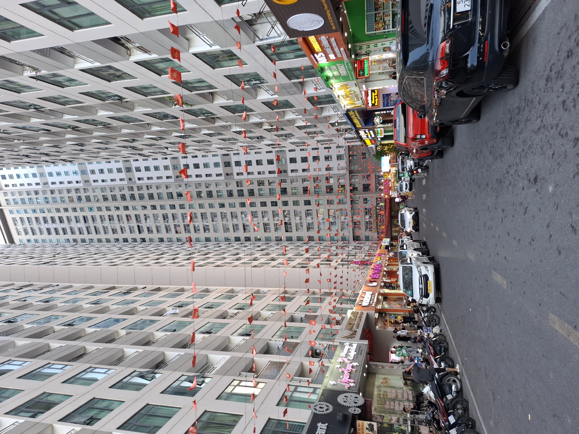 This image captures a street scene in Bac Nha Trang Ward, Vietnam, likely during daylight given the illumination. The foreground is dominated by a tall, modern building with numerous rectangular windows and balconies. Red flags are strung along the building's facade, creating a festive or celebratory atmosphere. To the right of the building, a street is lined with parked vehicles, including cars and numerous motorcycles. Shops and businesses with illuminated signs are visible along the street, suggesting a commercial area. One sign, partially obscured, reads "PHARMACY" and another displays "AN'S CONDOTEL". The general mood appears busy and lively, characteristic of an urban environment. The presence of parked motorcycles and people on the street indicates ongoing daily activity.