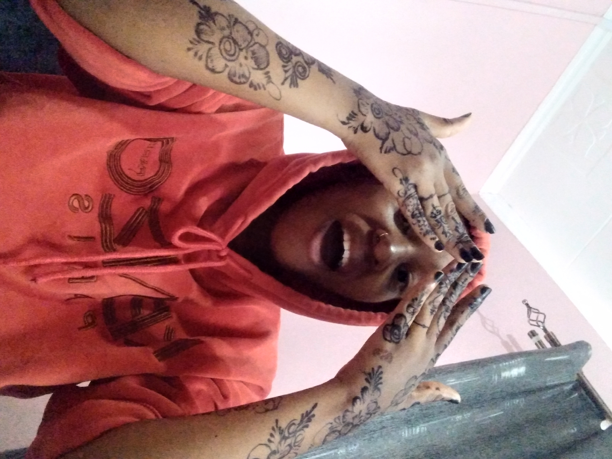 A young woman wearing an orange hooded sweatshirt poses with her arms raised, hands near her face, in an indoor setting. Elaborate henna-style tattoos are visible on her arms and hands. She has a nose piercing and black nail polish. The background includes a pink wall and a grayish-blue countertop or shelf with decorative items. The location is in the city of Unknown, Unknown.