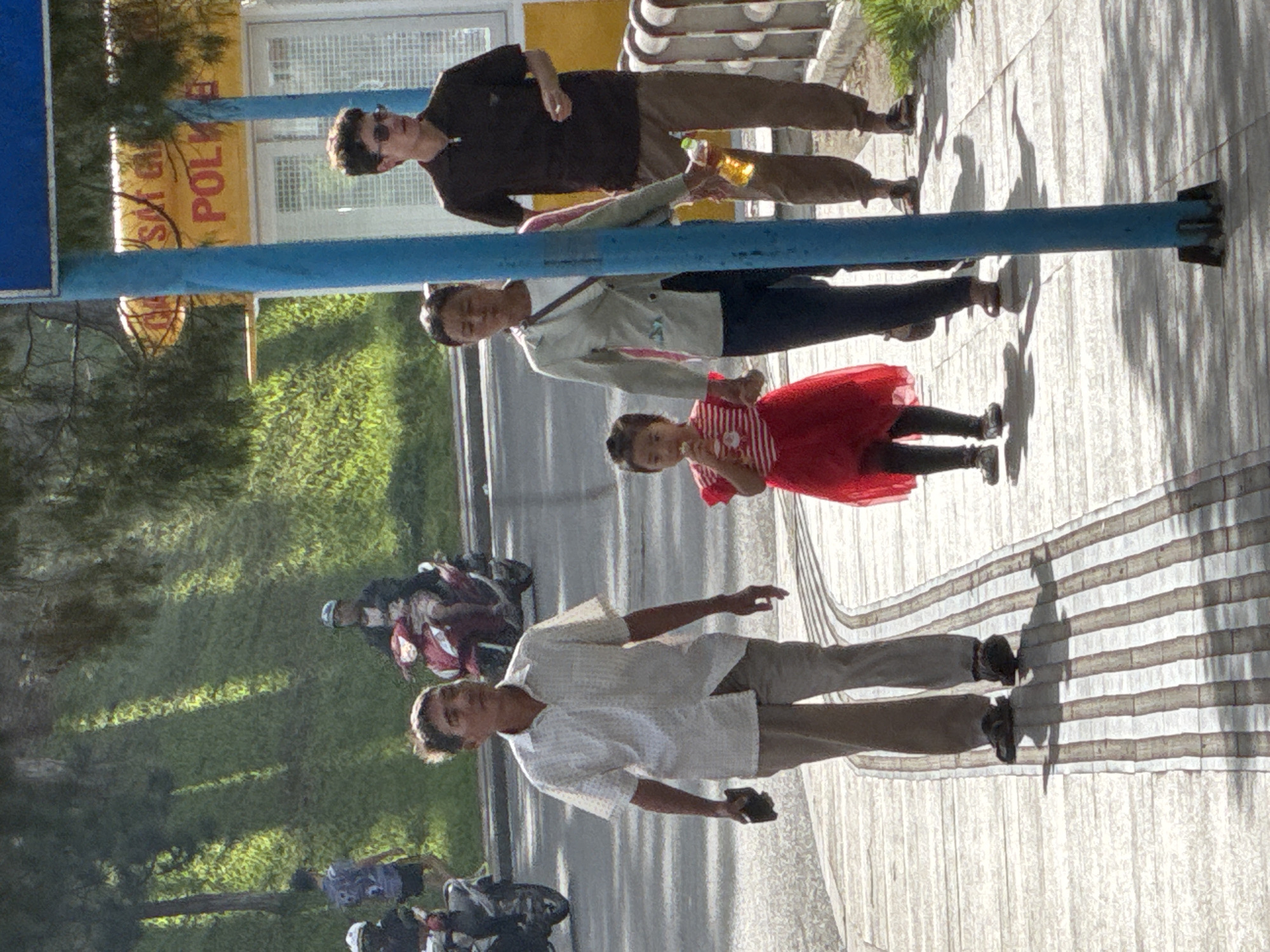 The image depicts a bright, sunny outdoor scene in Da Lat, Vietnam, featuring a sidewalk and a road with green, tree-covered hills in the background.
In the foreground, four individuals are walking towards the viewer on a light-colored sidewalk with darker parallel grooves. From left to right:
1. An adult male wearing a light-colored short-sleeved collared shirt, light trousers, and dark sandals. He holds a dark object in his left hand.
2. An adult female wearing a light-colored long-sleeved jacket over a white top, dark trousers, and dark shoes. She carries a dark shoulder bag and holds the hand of the young girl beside her.
3. A young girl, approximately 4-7 years old, wearing a red dress with a striped bodice and dark shoes. She holds a light-colored item to her mouth with her right hand and holds the woman's left hand.
4. An adult male, wearing a dark brown short-sleeved collared shirt, light brown trousers, dark sandals, and sunglasses. He holds a clear plastic bottle containing a yellowish liquid in his left hand. He walks alongside a low metal barrier and behind a prominent blue pole.
On the road in the midground, two motorcyclists are visible. To the left, a person in a white helmet and dark clothing rides a dark scooter away from the viewer. To the right, another person in a dark helmet and dark clothing rides a red scooter towards the viewer. Further back on a grassy slope, a person in a patterned dark blue t-shirt and dark shorts walks away from the viewer.
The background consists of lush green hills and trees. Above the right side of the sidewalk, a large yellow sign with red lettering is partially visible, displaying text that includes "SAY ĐA LẠT" and "POLKA". Strong sunlight creates distinct shadows from the people, the blue pole, and background trees across the sidewalk and grassy areas. Two prominent blue poles, possibly part of street furniture or a sign structure, stand on the sidewalk.