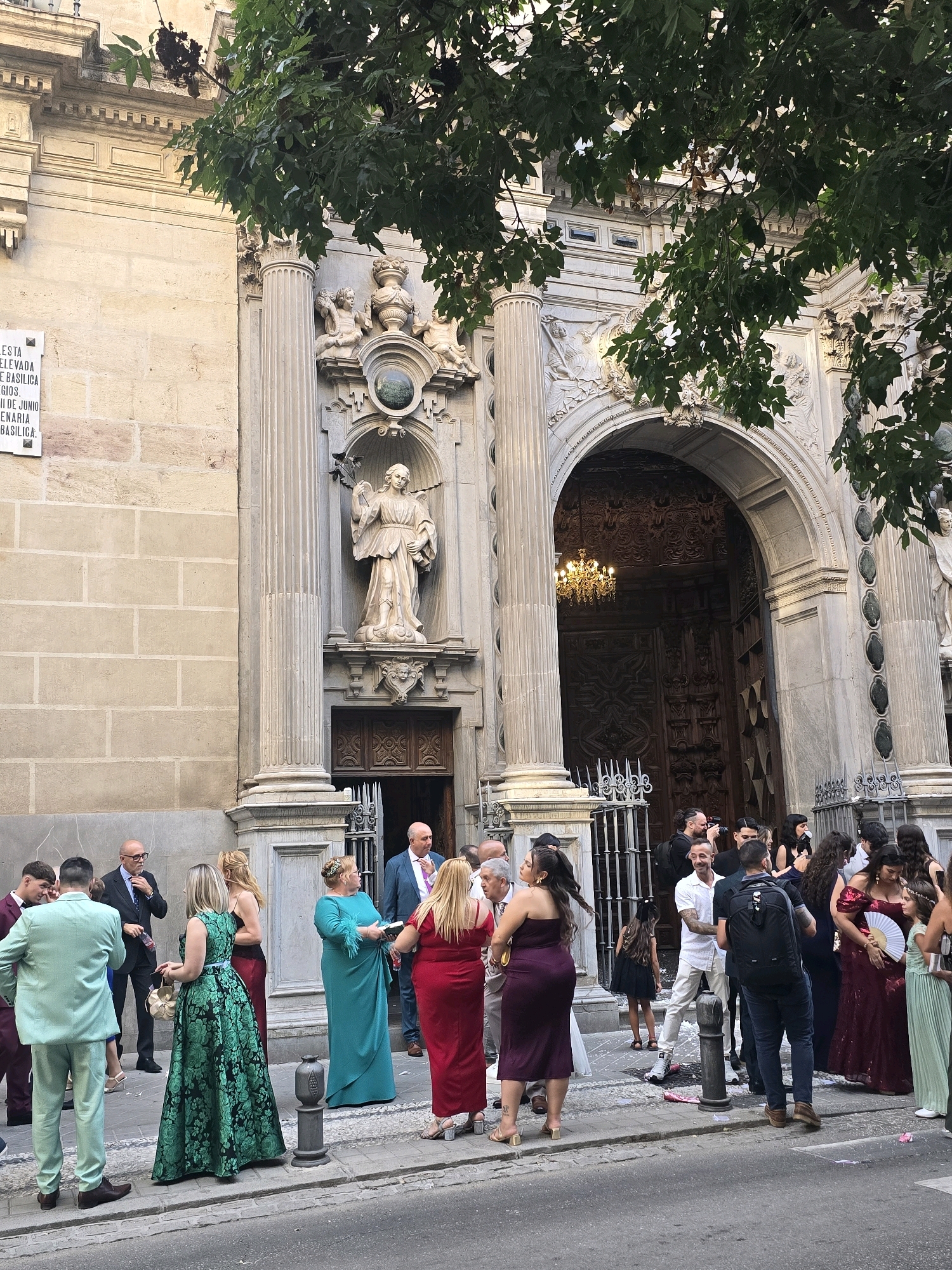 The image depicts a gathering of people outside a grand building, likely a church or cathedral, in Granada, Spain. The architectural style suggests a Baroque or Renaissance influence, with ornate stonework, tall columns, and a large arched doorway leading into the interior. A statue of a robed figure, possibly a saint, is prominently displayed in a niche above the entrance.
A group of individuals, dressed in formal attire, are standing on the cobblestone street and sidewalk. Several women are wearing long, elegant dresses in vibrant colors such as green, red, and teal. Men are mostly in suits or formal wear, with some in lighter colored suits. One man in a teal suit stands out in the foreground, with his back to the camera. Another man, in a burgundy suit, is to his left.
A cluster of people are gathered near the entrance, some appearing to be taking photographs. The atmosphere seems celebratory, possibly for a wedding or special event, as indicated by the formal dress and the presence of what looks like confetti or rose petals scattered on the ground in front of the doorway. Lush green foliage from trees overhangs the scene, partially obscuring the upper parts of the building. The light suggests it is daytime, with a slight shadow cast on the facade.