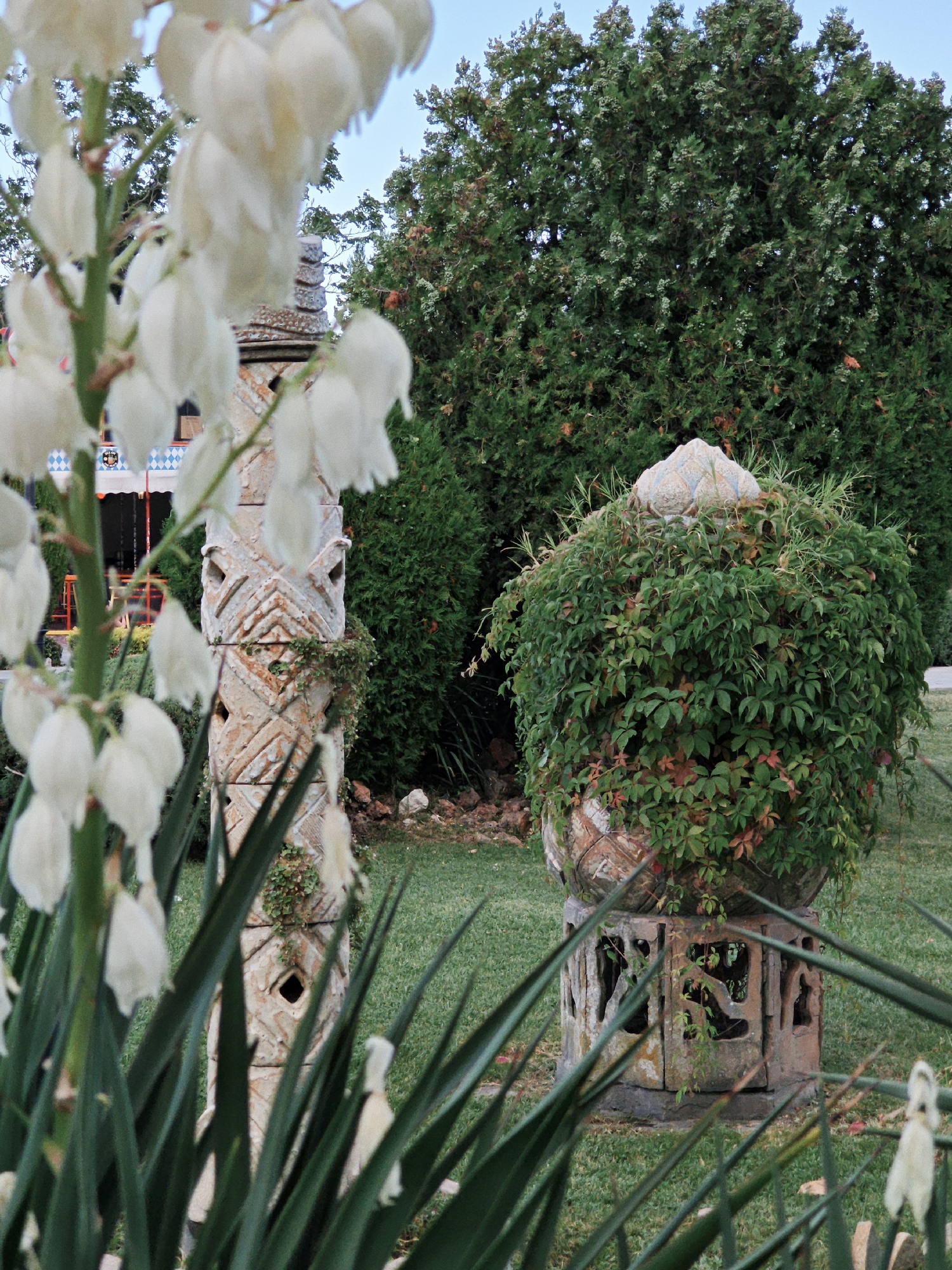 The image shows a garden scene with several ornate stone objects and lush greenery. In the foreground, a tall stalk of white flowers with bell-shaped blossoms is in soft focus, obscuring parts of the background. To the right of the flowers, a weathered stone column with intricate carvings and cut-out decorative shapes stands vertically. Further to the right, a large, round stone planter is overflowing with dense green ivy, with a tiered stone lid. Behind the planter, a massive, dark green tree with dense foliage dominates the upper portion of the image. The ground is covered in green grass, with patches of dry leaves and small rocks scattered around. In the far background, a building with a red awning and white and blue diamond-patterned fabric is partially visible. The sky above is a clear, pale blue.