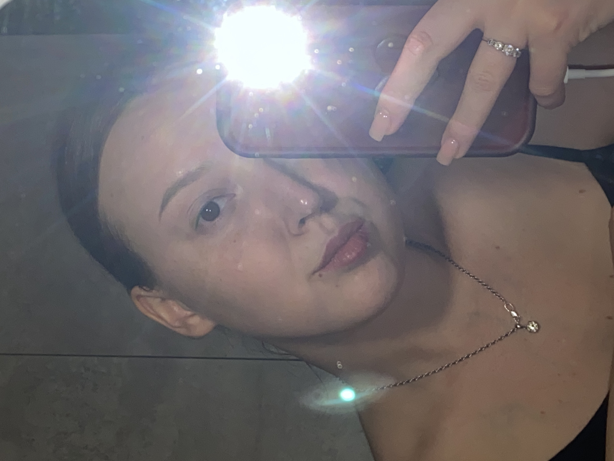 A person is taking a selfie in what appears to be a bathroom or tiled space. The person has fair skin, dark hair, and is wearing a silver necklace with a small pendant. They are holding a phone with a case, and a ring is visible on their finger. The flash from the phone is causing significant glare, obscuring some details. The location cannot be determined as Chaikovsky, Russia based on the image.