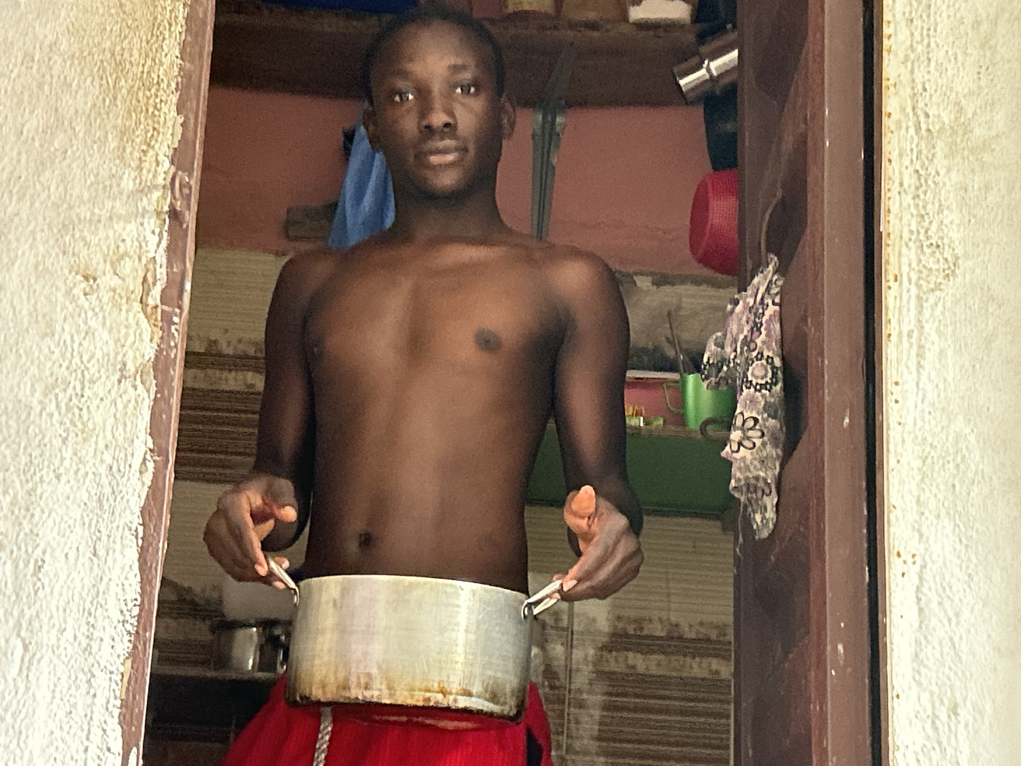 In a doorway in Ihiagwa, Nigeria, a young shirtless man stands. He is holding a large, metallic pot around his waist with both hands. His left hand grips the pot from the side, while his right hand holds a pair of small, metal tweezers, also gripping the pot. The man is looking directly at the camera with a neutral expression.
The background suggests an indoor setting, possibly a kitchen or living space, with shelves and various items visible. A blue cloth hangs on the left, and a red plastic container and a patterned cloth are on the shelves to the right. The walls are painted a pinkish hue, and the floor or lower walls have a striped pattern. The doorway has a wooden frame. The lighting suggests it is daytime, possibly midday, as there are no strong shadows indicating early morning or late evening. The overall impression is of a casual, everyday moment captured in a modest dwelling. There is no visible text in the image.