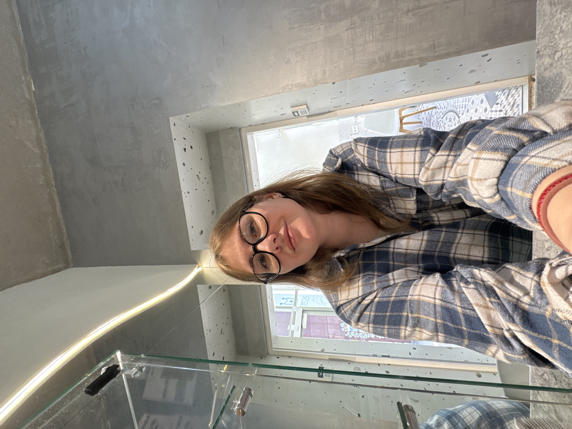 A young woman with brown hair and glasses, wearing a blue and white plaid shirt, poses for a selfie in what appears to be an indoor cafe or retail space. The background features a concrete wall, a glass partition, and a doorway leading to an area with patterned flooring and a white chair. A strip of warm LED lighting runs along the ceiling. The location is identified as being in the city of Perm, Russia.