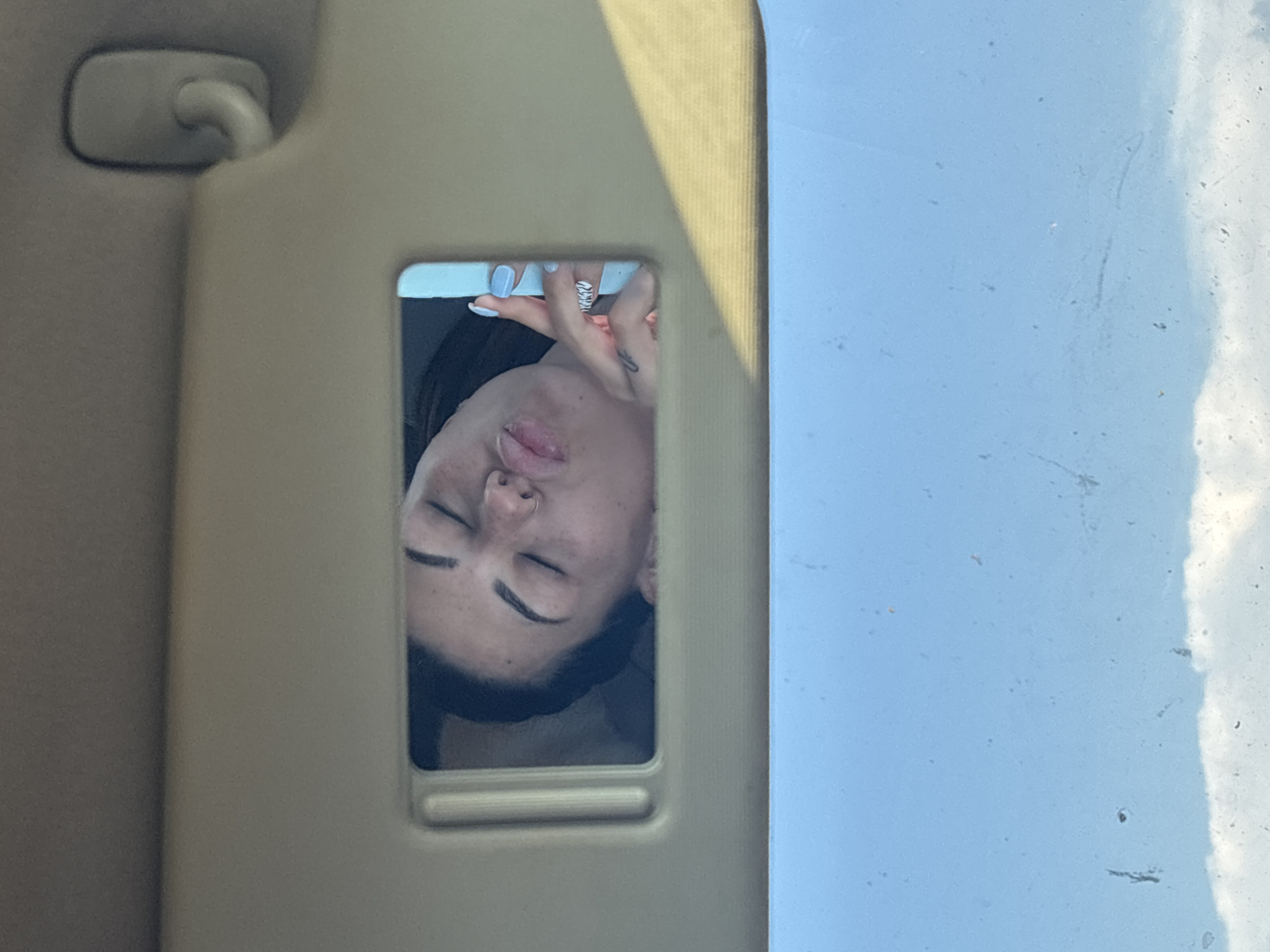The image shows a person's face reflected in a car's visor mirror. The person appears to be a young woman with closed eyes and her tongue slightly out. She has light skin, dark eyebrows, and a nose ring. Her hand is visible, with light blue nail polish and a ring on one finger, suggesting she is taking a selfie or adjusting her appearance. The reflection is in a vertical orientation, indicating the photo may have been taken from an unusual angle or the original image was rotated. The background of the mirror frame is a light beige color, typical of a car's interior. Beyond the visor, a blue, cloudy sky is visible, suggesting it is daytime and likely fair weather. The setting is inside a car, likely in Sofia, Bulgaria. No text is visible in the image. The overall impression is a candid moment captured while in a vehicle.