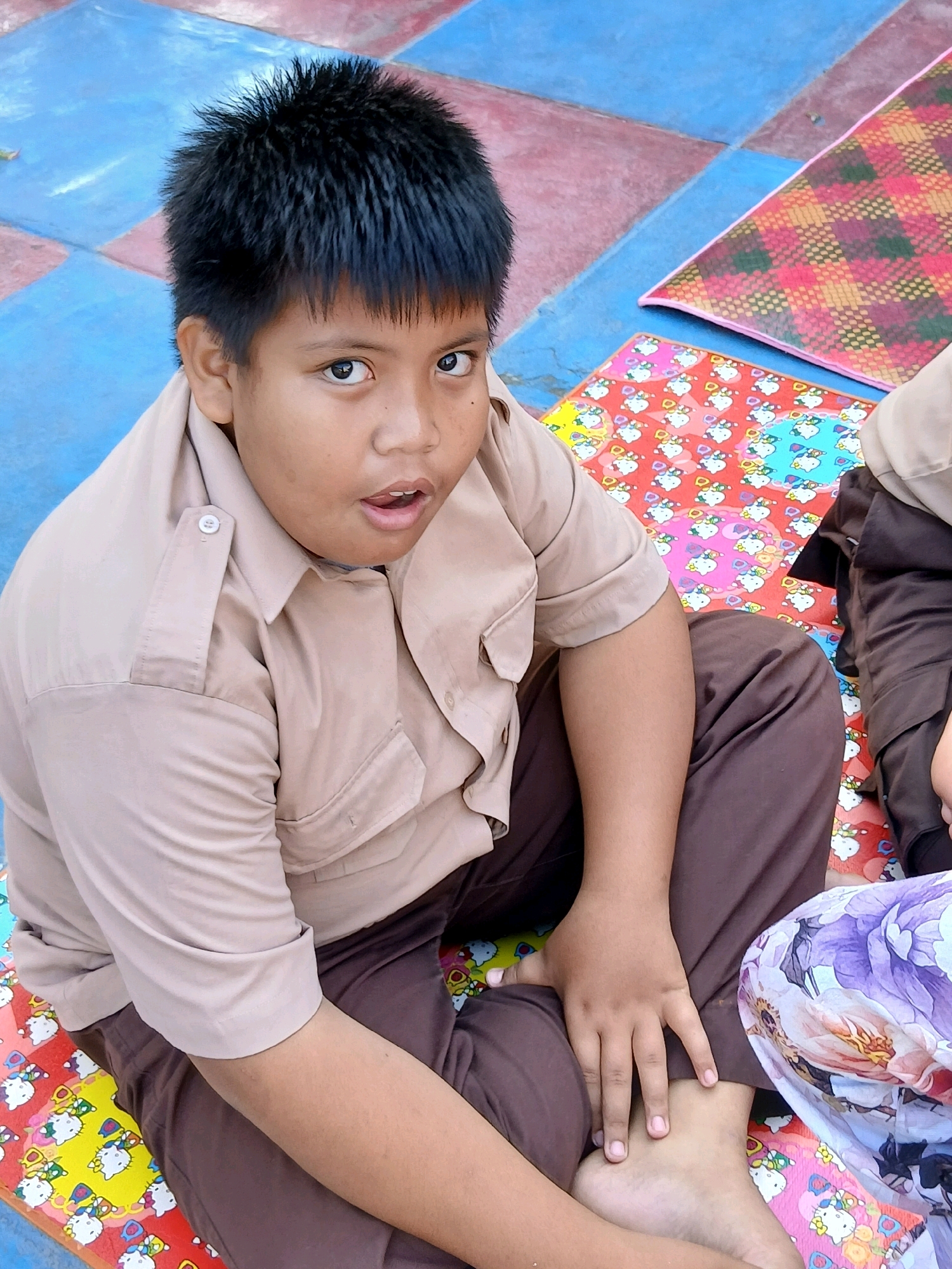 A young boy, likely of Indonesian ethnicity, is seated on a colorful mat with a checkered background in what appears to be an outdoor or semi-outdoor setting. He is wearing a light brown uniform shirt with buttons on the collar and pockets, and dark brown pants. His hair is dark and spiked. The mat features a repeating pattern of cartoon characters, possibly Hello Kitty, in various colors including red, yellow, and blue. Another mat with a pink, yellow, and green geometric pattern is partially visible to his right. His left hand rests on his leg, and his feet are bare. The overall scene suggests a casual gathering or activity, possibly at a school or community event in Gorontalo, Indonesia.