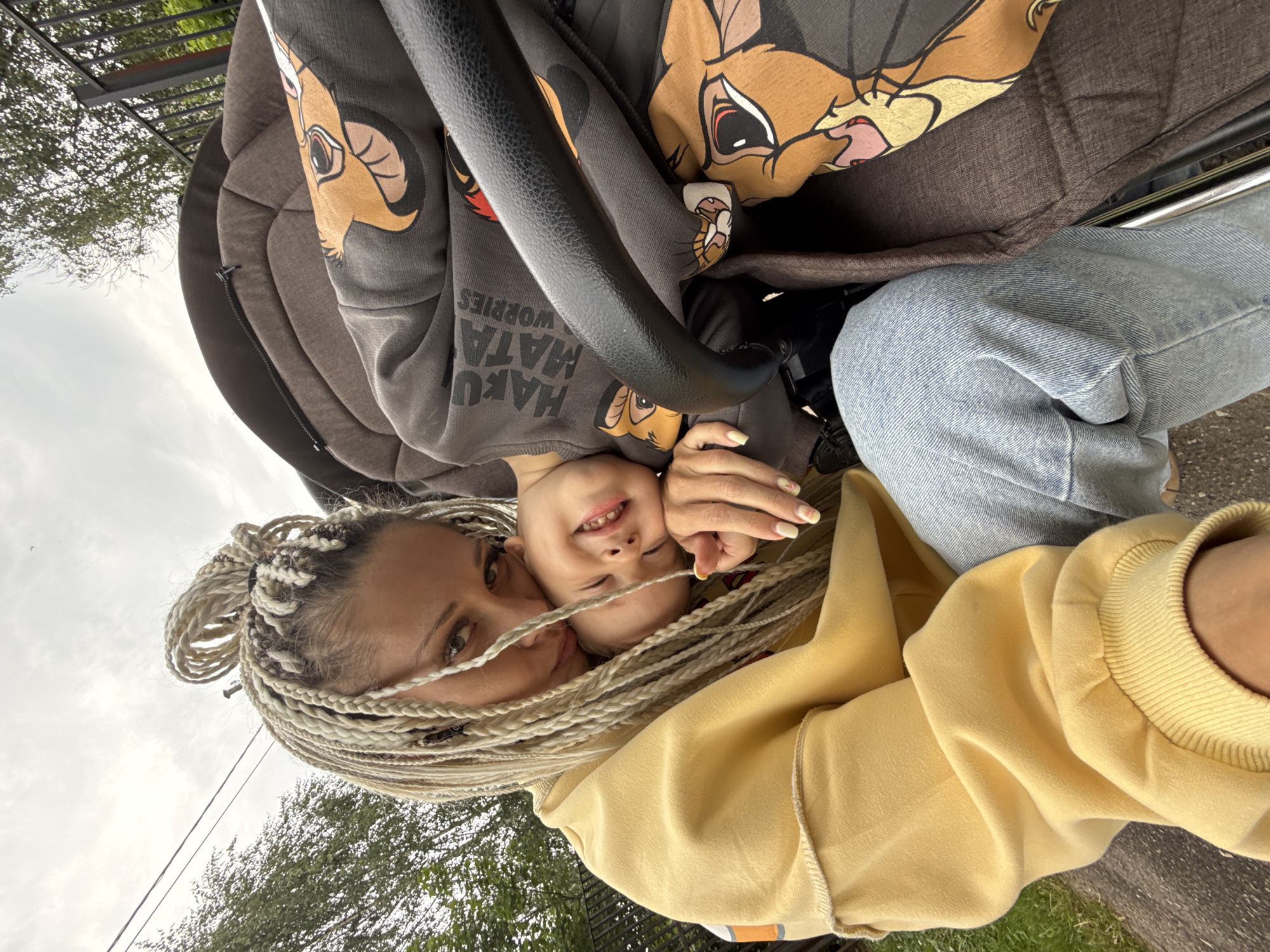 In Rusanovo, Russia, a blonde woman with elaborate braided hair is leaning forward and embracing a smiling baby in a stroller. The woman is wearing a yellow sweatshirt and blue jeans. The baby is wearing a dark gray sweatshirt with a Lion King graphic. The stroller is dark brown. The background consists of trees, a fence, and an overcast sky. The photo was taken outdoors.