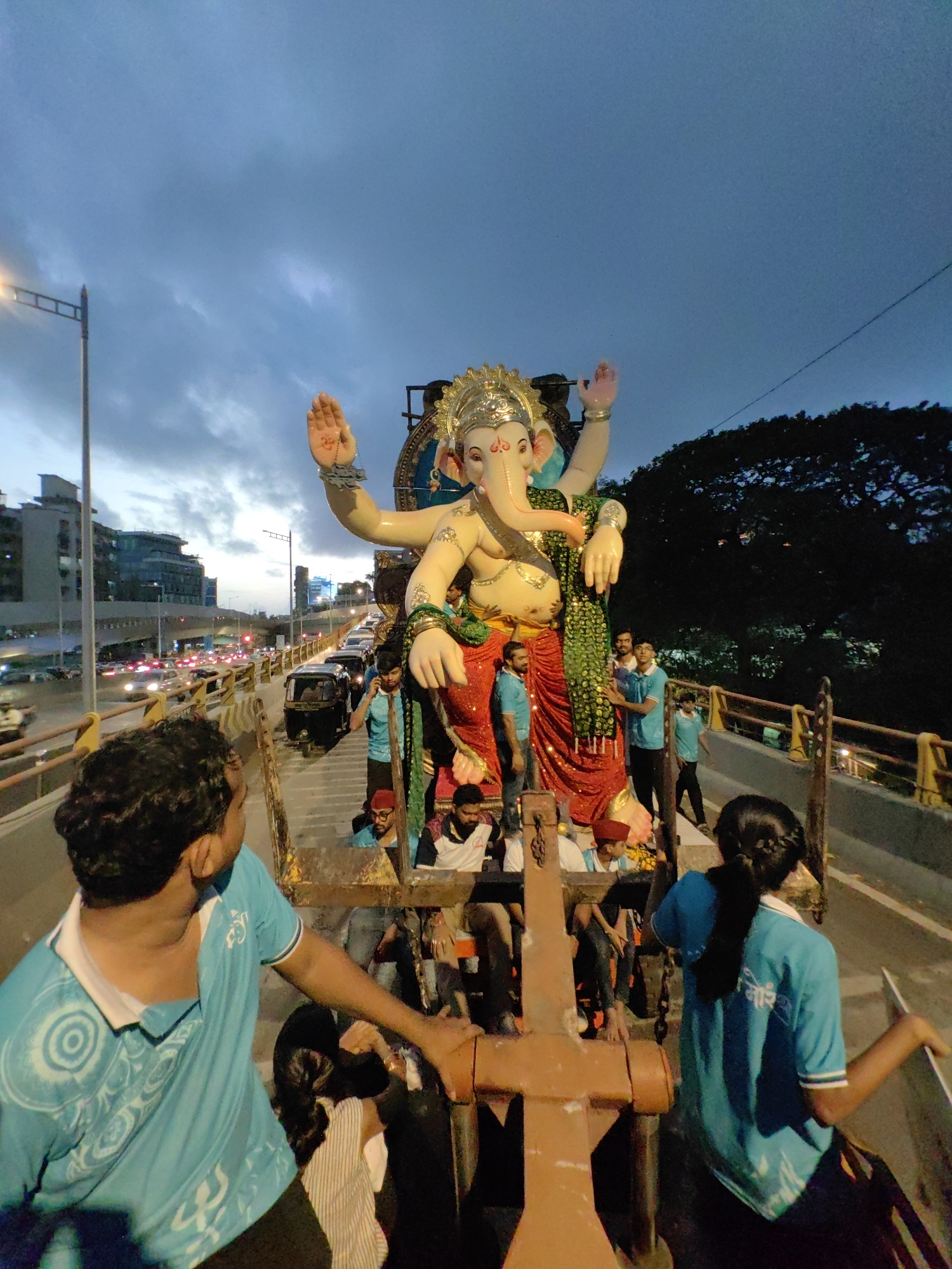 The image depicts a large statue of Lord Ganesha being transported on a truck, likely as part of a religious procession in Mumbai, India. The statue is vibrant, with multiple arms and adorned in traditional attire. A crowd of people, mostly young men and some women, are visible around the statue, some wearing blue t-shirts with an emblem. The truck is on a road, with traffic visible in the background, including cars, auto-rickshaws, and streetlights, suggesting an urban setting in Mumbai. The sky is overcast, indicating either dusk or dawn. The scene captures a moment of celebration and community involvement in a religious festival, possibly Ganesh Chaturthi.