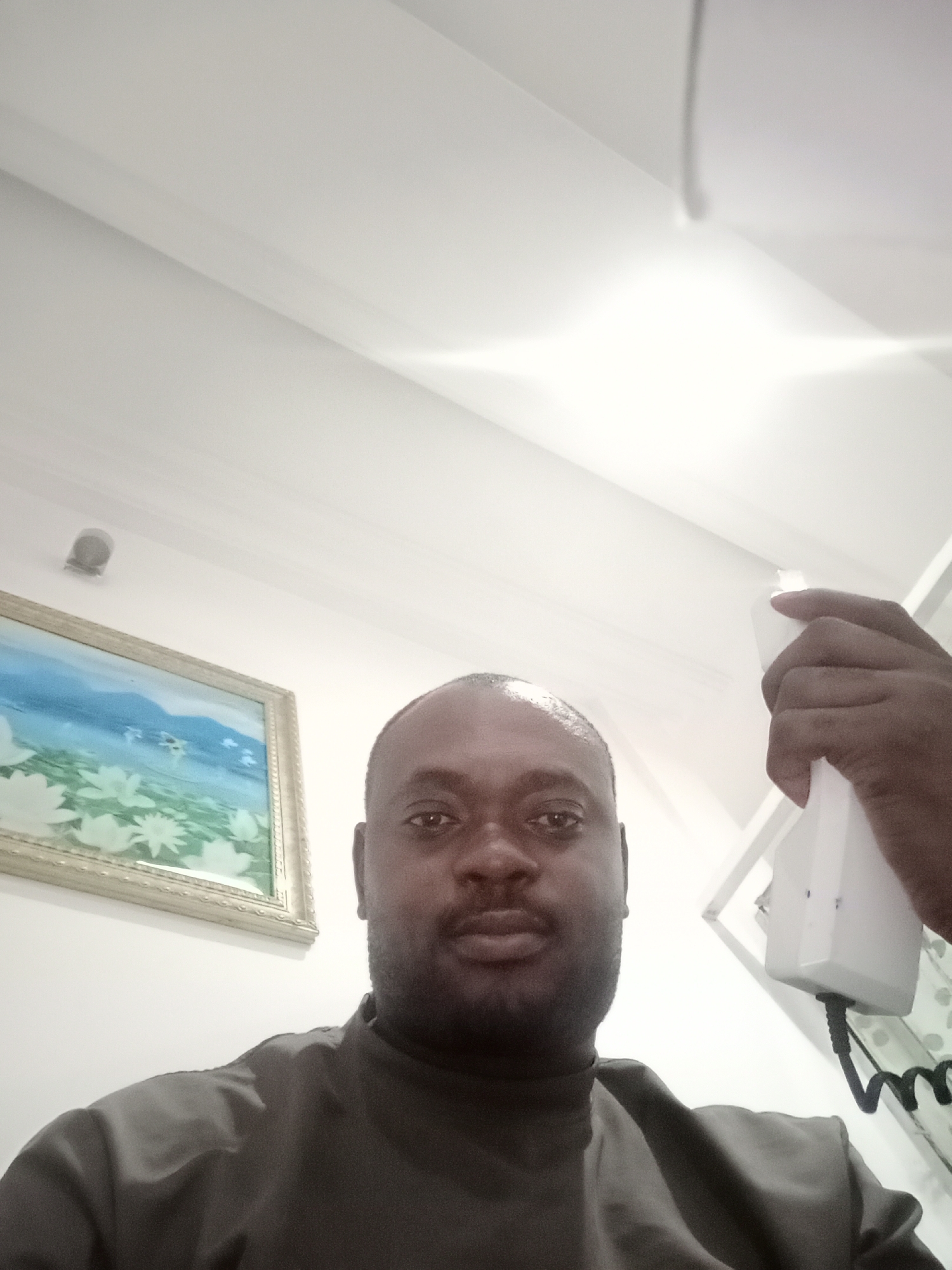 A dark-skinned man is holding a white landline telephone receiver near his right ear. He is facing the camera. He is wearing a dark collared shirt. Behind him is a white wall. There is a framed picture on the wall to his left. The ceiling is white, and there appears to be a light fixture above. The man is located in Gwarinpa, Nigeria.