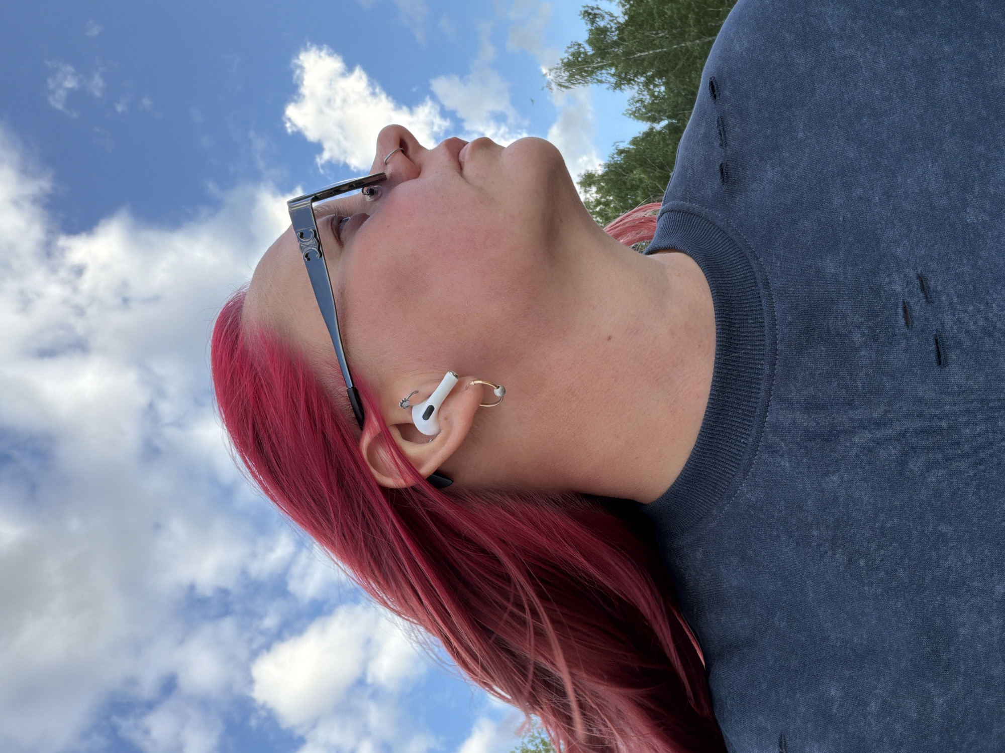 An outdoor, eye-level, medium close-up profile shot shows a woman with vibrant pink hair, wearing a dark, distressed crewneck sweatshirt and eyeglasses. She has a hoop earring in her left ear and an earbud. The woman's gaze is directed upwards and to the right, against a backdrop of a bright blue sky with scattered white clouds. In the distance, a line of green trees is visible. This scene is likely in Chaikovsky, Russia, given the context of the provided metadata.