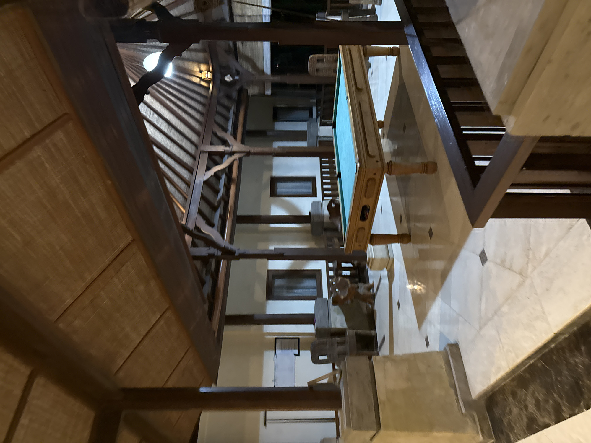 The image shows an indoor scene with wooden architecture and marble floors. The perspective is from a high vantage point looking down. A pool table with a green felt surface is a prominent object in the middle of the frame. It has wooden legs and a decorative wooden frame. The surrounding area appears to be a spacious, open-plan room, possibly a lobby or lounge area. Wooden beams and intricate wooden carvings are visible, suggesting a traditional or rustic design. There are windows with dark frames, and some lighting fixtures are visible, including a wall-mounted lamp. The floor is tiled with large, light-colored marble tiles, some of which have a darker diamond pattern. The time of day appears to be evening or night, as the lighting is dim and artificial. There are no people visible in the image. The setting suggests a hotel, resort, or a large residential building in Kuta Selatan, Indonesia, given the location context. The overall impression is one of a well-maintained, possibly luxurious, interior space with a focus on natural materials and craftsmanship.