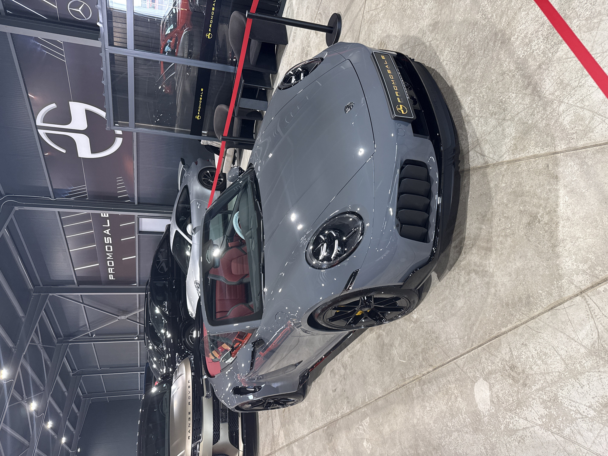 A grey convertible sports car with a red interior is parked on a concrete floor inside a building. The car has black rims and yellow brake calipers. In the background, several other cars are visible, including a gold Range Rover and a silver Porsche. A large banner with a logo and the word "PROMOSALE" is displayed on the wall. There are also two black chairs and a small table set up in front of a glass partition. Red tape is stretched across the foreground. The location is PROMOSALE in Plovdiv, Bulgaria.