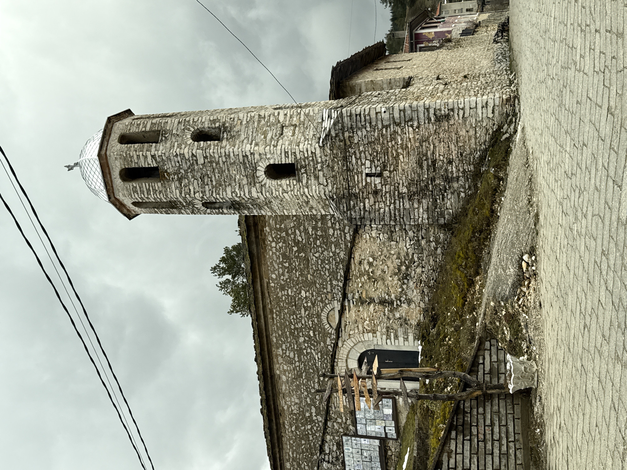 The image shows the Church of St. Theodore Tyron and Theodore Stratelates, located in Lilkovo, Bulgaria. The stone building consists of a tall tower with arched windows topped with a metal dome, and a lower section. A doorway is visible on the lower section, with a wooden structure placed in front of it. A paved walkway is to the right. Above the building are power lines against a cloudy sky.