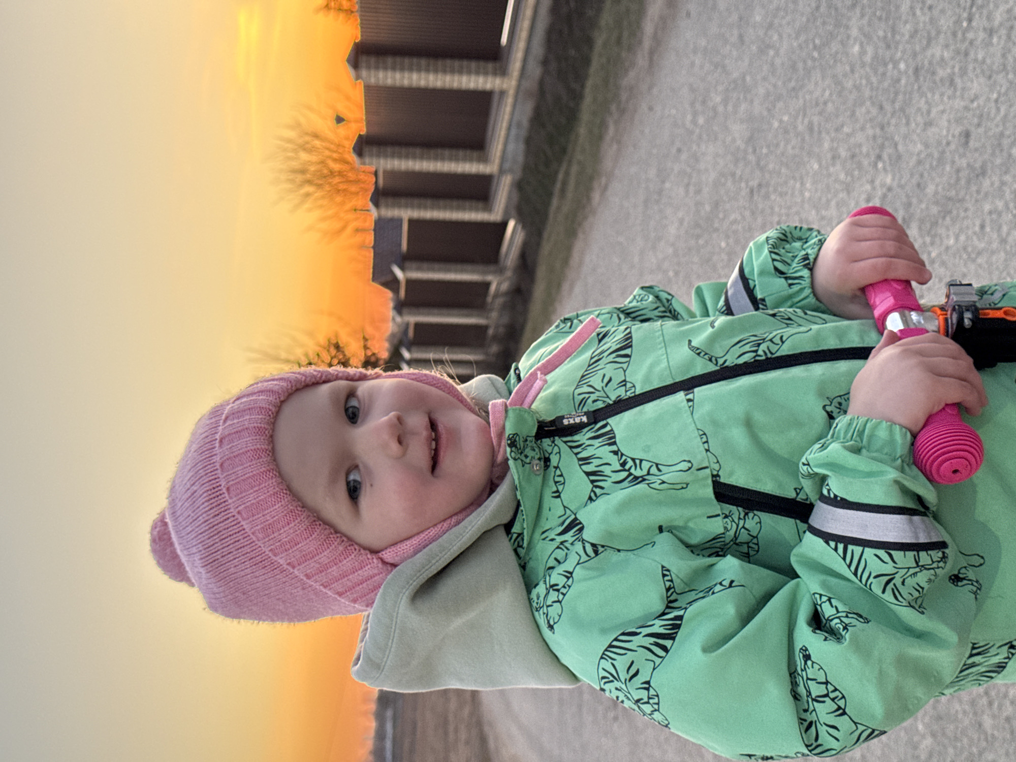 A young child with a pink knit cap and a green jacket patterned with black tigers is standing on an asphalt surface outdoors. The child is holding a pink object, potentially part of a scooter or toy. Behind the child is a building with horizontal architectural details, and above it, the sky is a gradient of orange and yellow, indicating sunrise or sunset. The photograph appears to have been taken in Горный, Russia, based on provided location context.