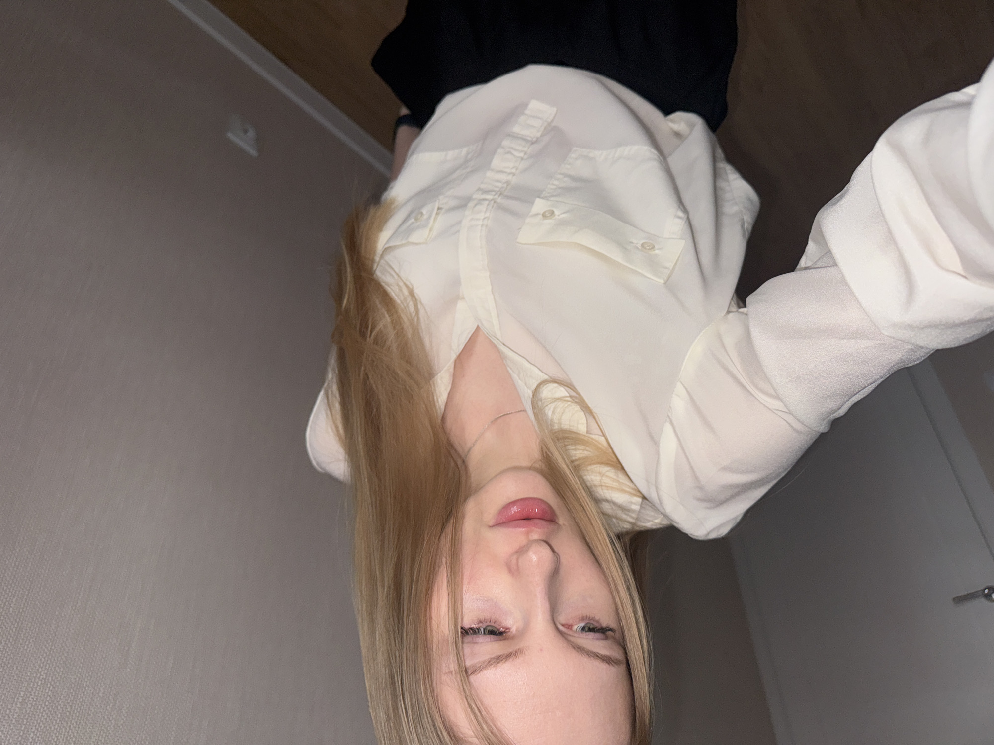 A woman is seen from a low angle, taking a selfie. She has long blonde hair, fair skin, and is wearing a light-colored blouse and a dark skirt or shorts. The background shows a plain wall. The exact location in Perm, Russia, cannot be determined from the image.
