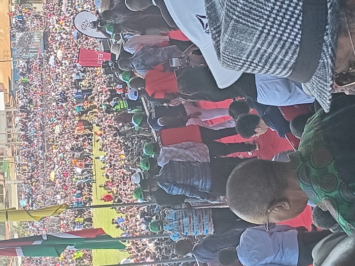 This image captures a large-scale outdoor public gathering or rally in Siaya, Kenya, under clear, bright daylight. The scene is densely populated with a vast crowd filling a stadium or expansive open field, extending far into the background.
In the foreground, on the left, a Kenyan flag (featuring black, red, and green stripes with a Maasai shield) is prominently displayed alongside a yellow flag. On the right, the backs of several individuals are visible. One prominent figure has a bald head and wears eyeglasses, dressed in a green garment with circular patterns. Nearby, another person wears a distinctive black and white checkered jacket. A white shirt or banner featuring the "Reebok" logo is also visible in the upper right.
The midground features a red-carpeted area or platform where key individuals are gathered. Many in this area are wearing matching green caps or hats and some are in red shirts, suggesting a unified group or supporters. Others are seen in light blue shirts. Behind this platform, a red banner with the white text "DTB" is clearly visible, along with a white sign bearing a unique logo (resembling a snowflake or star).
The sheer number of people in the background, tightly packed into the stands and across the field, indicates a significant event. Some members of the crowd are holding umbrellas, likely for sun protection. The overall atmosphere suggests a major public event, possibly a political rally, community celebration, or public address, characterized by widespread participation.