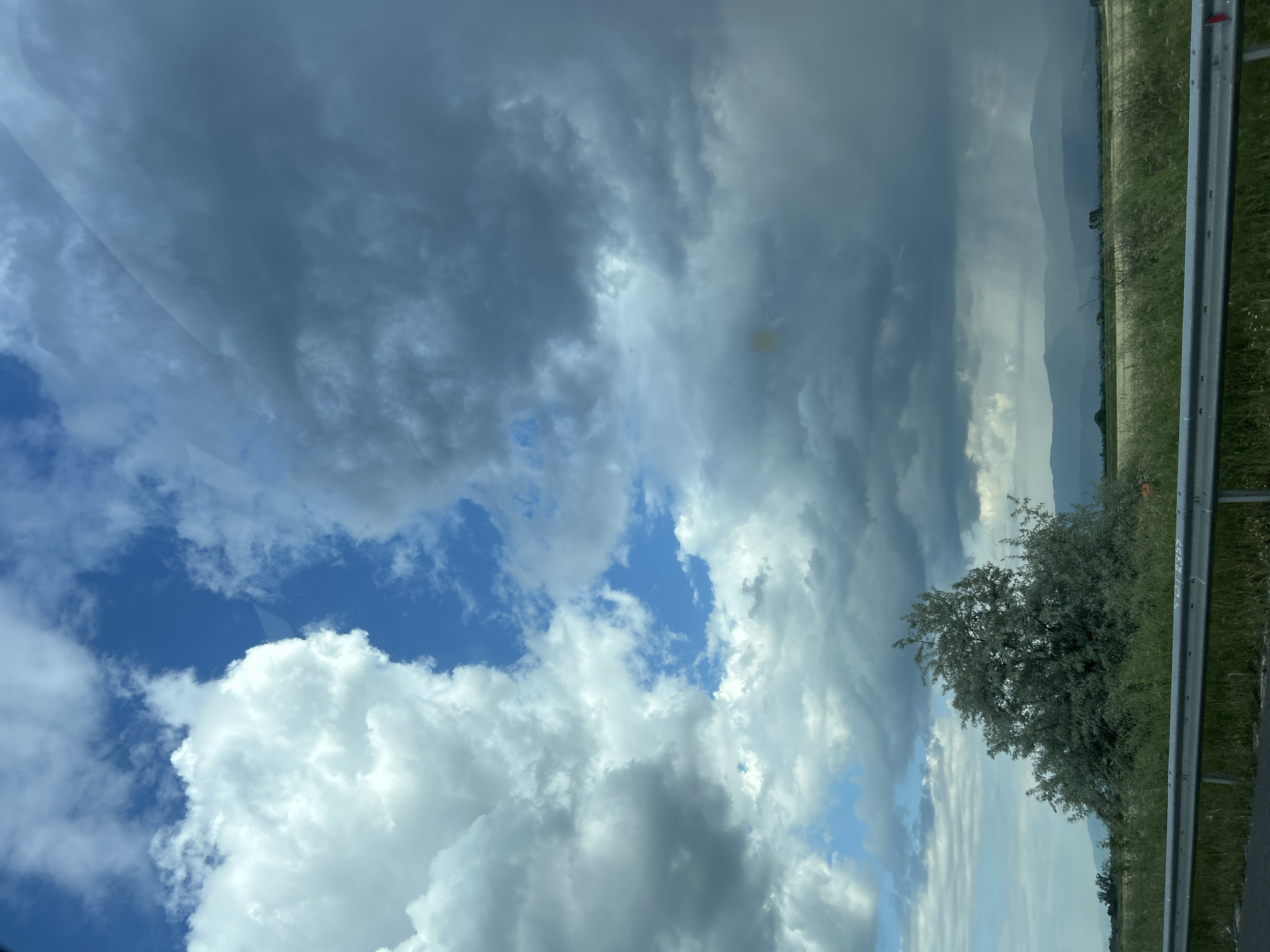 The image depicts a view from a vehicle, likely a car, in the vicinity of Malo Konare, Bulgaria. The focus is on the sky, which features a mix of dark, ominous storm clouds and patches of bright blue sky with white, puffy clouds. A strip of land is visible along the right side of the frame. This includes green vegetation, possibly a field or roadside area, a line of trees, and distant, hazy mountains. A grey metal guardrail is visible near the right edge, partially obscuring the vegetation. A small section of a tree is visible near the guardrail. The lighting suggests it is daytime, but potentially overcast due to the storm clouds. There is no indication of people or interactions within the scene.