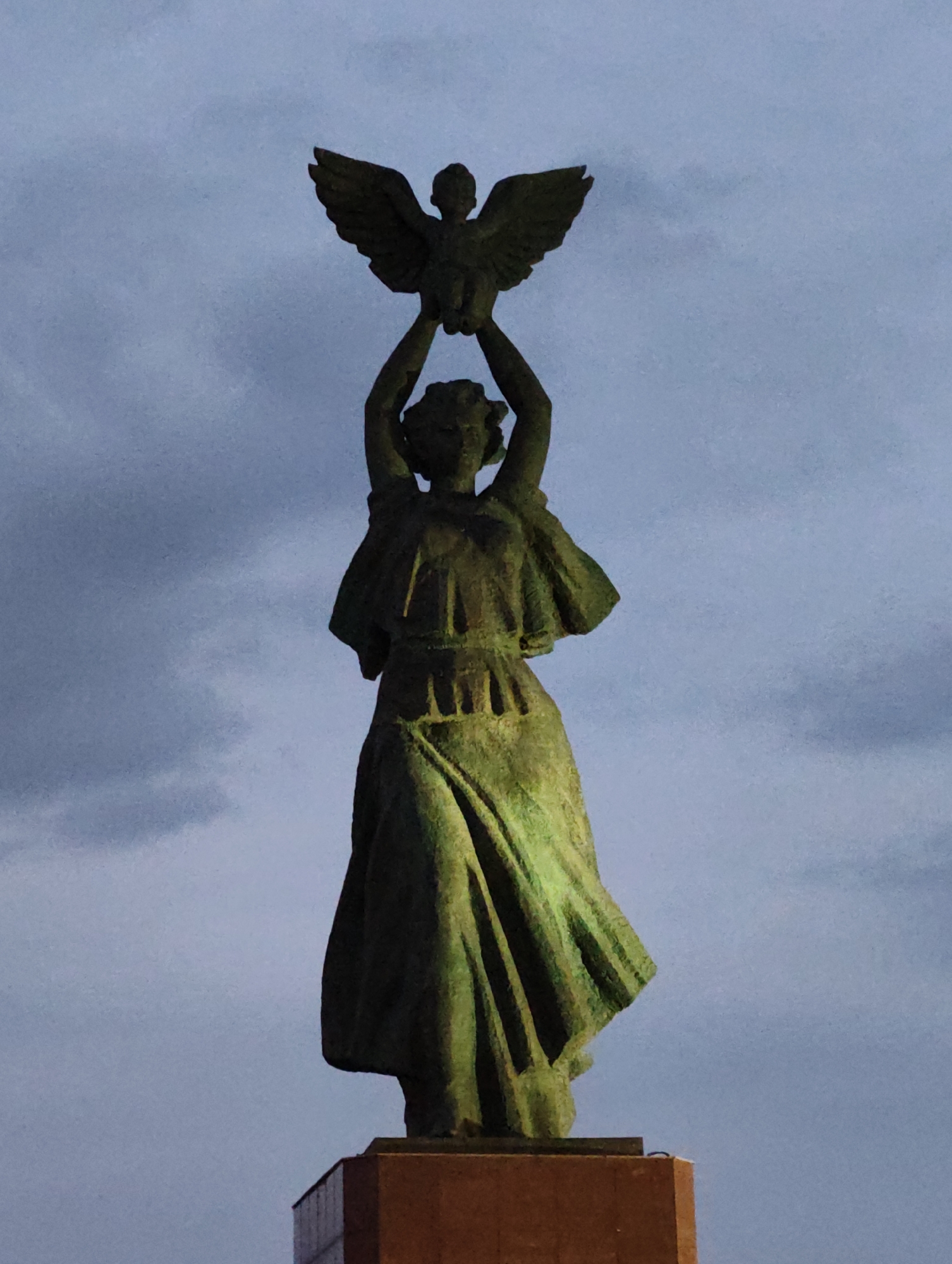 A weathered bronze statue of a female figure stands atop a reddish-brown pedestal against a cloudy, twilight sky. The figure is depicted holding a bird aloft with both hands, its wings spread. The statue's patina suggests age, with green and dark brown tones visible on its surface. The female figure wears a flowing, floor-length gown with folds and drapery. The statue is located in the city of Aihui, China. The overall scene is somber and majestic.