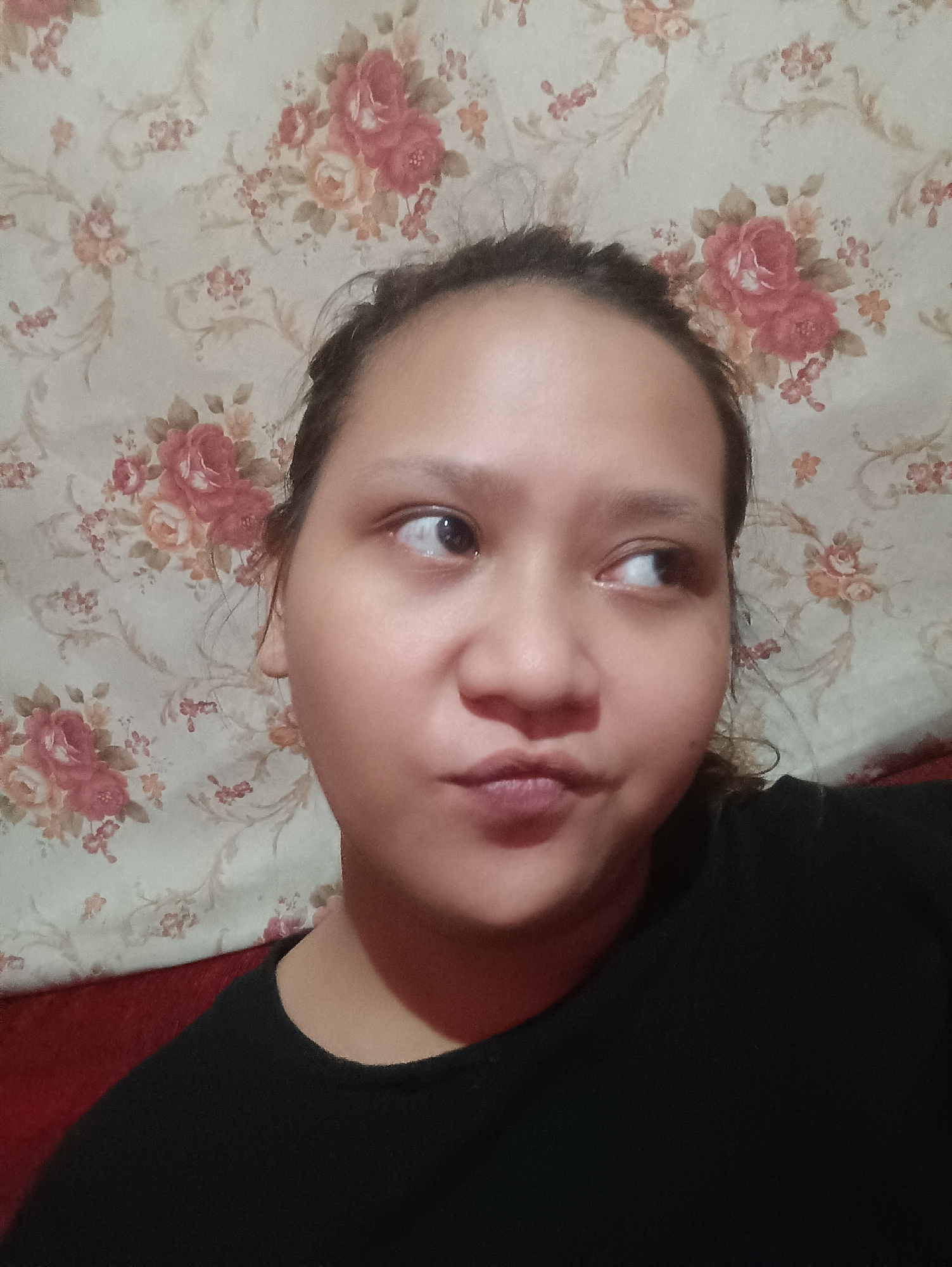 A close-up, head-and-shoulders shot shows a young woman in Depok, Indonesia, looking to her right with her lips pursed. She is wearing a black t-shirt, and her hair is pulled back. The background is a floral-patterned wallpaper with roses and other flowers in pink and peach hues. A red sofa is partially visible in the lower left corner. The lighting is dim, creating a soft focus effect. The image appears to be a selfie, taken indoors.