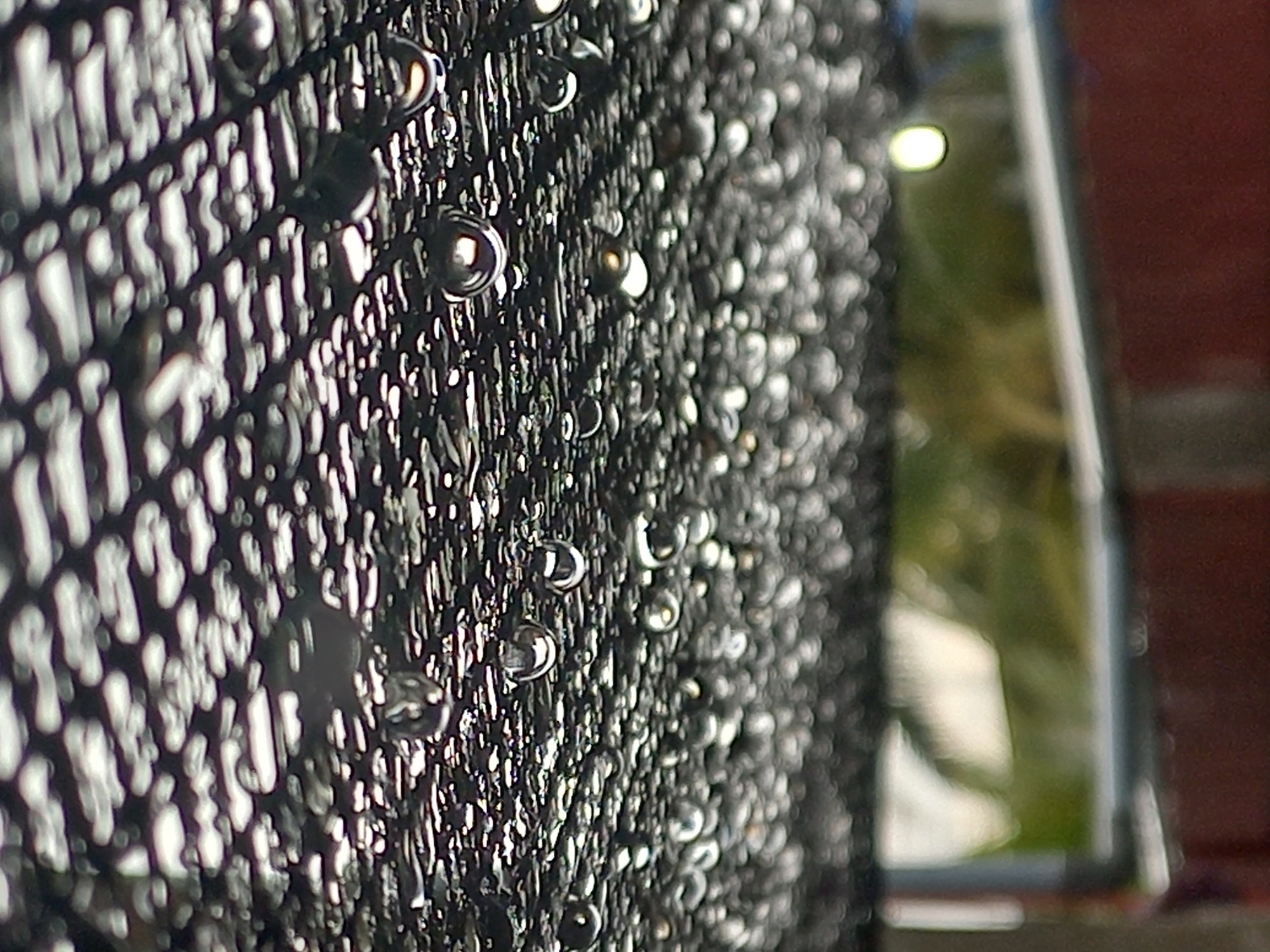 The image displays a close-up, macro-style shot of a dark, textured mesh or screen densely covered in numerous water droplets. The mesh exhibits a distinct, regular grid-like pattern. The water droplets vary in size and shape, clinging to the surface, with many appearing spherical and translucent, reflecting bright highlights from an unseen light source. These reflections create a sense of depth and luminosity.
The foreground and midground, encompassing the mesh and water droplets, are in sharp focus. The background is heavily blurred with a shallow depth of field. Indistinct patches of green, suggestive of foliage or grass, and a reddish-brown area, possibly a brick wall or another architectural structure, are visible in the background. A bright, circular light reflection or source is discernible within the blurred area, alongside a prominent vertical line that could be an edge or frame, such as a window or door frame.
The presence of water droplets indicates conditions such as recent rain, dew, or condensation. The lighting appears to be natural daylight, given the clear reflections. The scene suggests an outdoor setting, possibly a window screen, a fence, or an architectural detail, located in Puloblang, Indonesia. There are no people, animals, or visible text in the image.