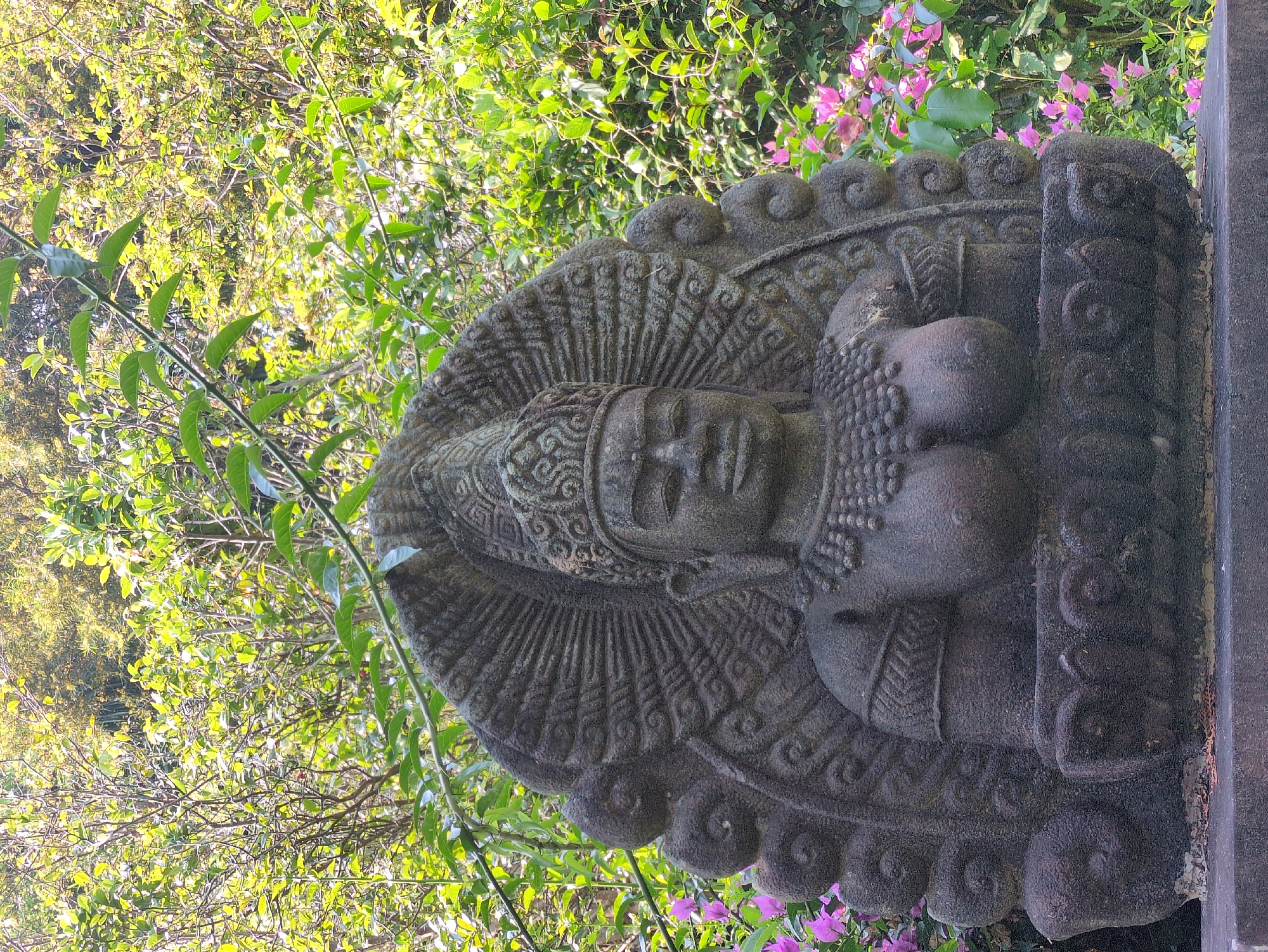 A stone sculpture rests in an outdoor setting, likely a garden or park. The sculpture appears to be a deity, perhaps a Buddha or Hindu figure, depicted with elaborate headdress, necklace, and ornamentation. The sculpture is made of dark, rough-hewn stone. The background features lush foliage, including green bushes and pink flowering plants. There is no evidence that this photograph was taken in Tuy Hòa, Vietnam.