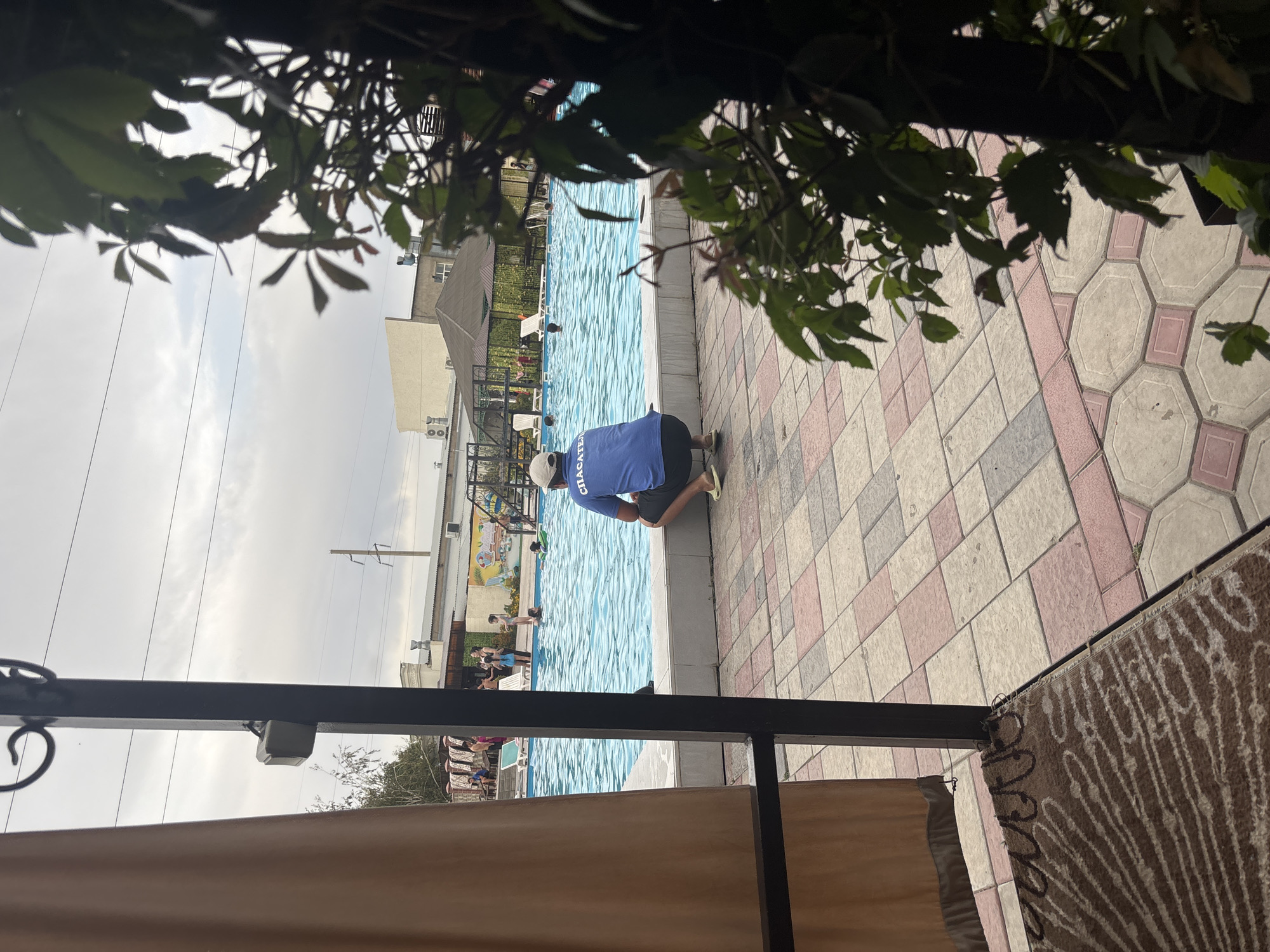 A man wearing a blue shirt with the word "СПАСАТЕЛЬ" (Rescuer) and dark shorts crouches on the edge of a swimming pool. The pool is filled with clear blue water and is surrounded by a tiled walkway. In the background, several people, including children, are in and around the pool. The scene appears to be an outdoor recreational facility. The architectural style of the buildings in the background, with their flat roofs and visible air conditioning units, suggests a climate common in Central Asia. The presence of utility poles and overhead wires further situates the location within a developed urban or semi-urban area. The foreground features a patterned mat and a draped curtain, indicating a covered or partially enclosed viewing area overlooking the pool. The sky is overcast, suggesting cloudy weather.