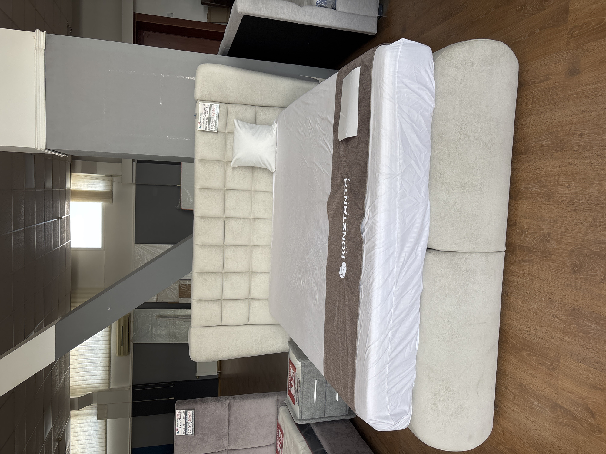 This is a high-angle, rotated shot of a bed in what appears to be a furniture showroom. The bed is upholstered in a light-colored, tufted fabric with a white duvet and a brown runner. A small white pillow rests at the head of the bed. To the right of the bed is a rounded, light-colored ottoman or bench. The floor is made of polished wooden planks. In the background, there are glimpses of other furniture, shelving units, and a window with blinds. There is a price tag on the headboard of the bed with handwritten text and a printed label. Another price tag is visible on a gray storage box near the foot of the bed. The overall lighting suggests it is daytime, likely indoors. The scene is static, with no people or activity depicted. Visible text includes "KONSTANTА" on the runner of the bed and handwritten numbers and Cyrillic text on the price tags, such as "43400" and "36.900" with what appears to be "грн" (Hryvnia, Ukrainian currency). The location context provided is Zaporizhzhia, Ukraine.