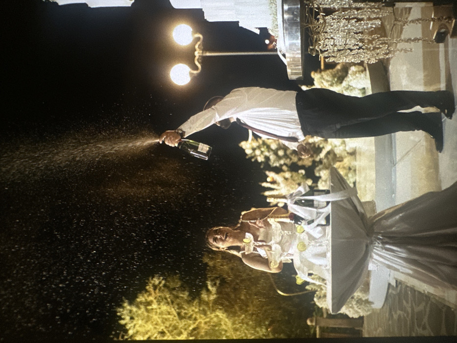 This image captures an outdoor nighttime event in Stains, France. A man, positioned on the right, wears a white long-sleeved shirt, a dark tie, and dark trousers. He holds a champagne bottle, identified as Moët & Chandon, with his right arm raised, spraying liquid upwards and to the left. To his left, a woman in a light-colored, possibly white, sleeveless dress stands smiling and looking upwards, appearing to react to the spraying champagne. Between the two individuals, a circular table draped in a light, reflective fabric (likely silver or white) holds several items. On this table, multiple champagne coupes are arranged in a tiered formation, each garnished with a lime wedge. Two additional champagne bottles, wrapped with white ribbons, are also present on the table. To the far right, a multi-tiered white cake on a silver stand, featuring ornate crystal-like dangling decorations, is partially visible. The background reveals illuminated trees to the left and a double-globe street lamp behind the man. The ground appears to be a paved surface. The sky is dark, indicating a nocturnal setting, with artificial lighting illuminating the scene.