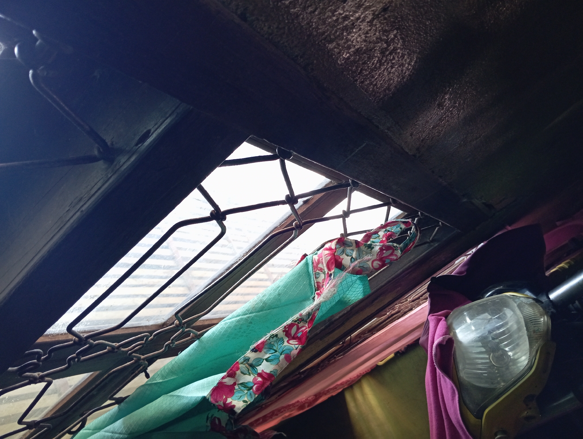 This is a close-up, eye-level, medium shot of a motorcycle's headlight, partially covered by a pink cloth. The headlight is yellow and has a clear, ribbed lens. Behind the motorcycle, a turquoise fabric with a floral pattern of pink and blue flowers is visible, draped over a metal window grate. The window itself is partially obscured by the grate, and appears to look out onto a bright, overcast sky. To the right of the window, a dark, textured wall is present. The scene suggests an indoor setting, possibly a garage or covered outdoor area, in Rejosari, Indonesia.