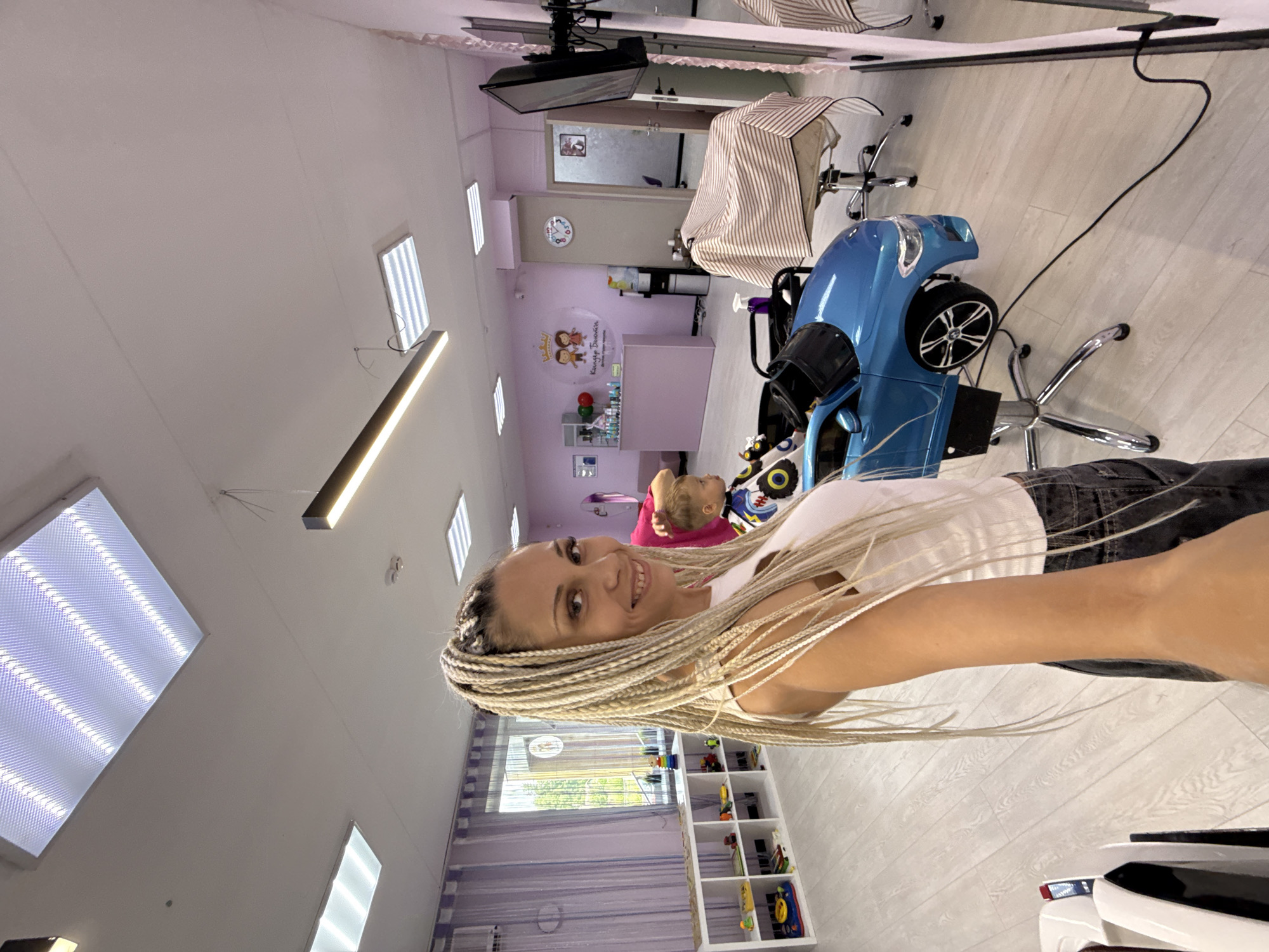 The image captures a well-lit interior of what appears to be a children's hair salon, possibly located in Rusanovo, Russia. A woman with blonde braided hair, wearing a white tank top and dark jeans, is taking a selfie, smiling at the camera. In the background, a young boy, dressed in a barber's cape with a colorful pattern, sits in a blue toy car that functions as a child's salon chair. The salon features light-colored wooden flooring and light pink walls adorned with decorations. To the left, a white shelving unit is filled with toys and children's books. A reception desk with a decorative logo that reads "Королевский Бомонд" (Royal Bohemian) is visible, along with a water cooler and a clock on the wall. The overall environment is clean, organized, and designed to be child-friendly.