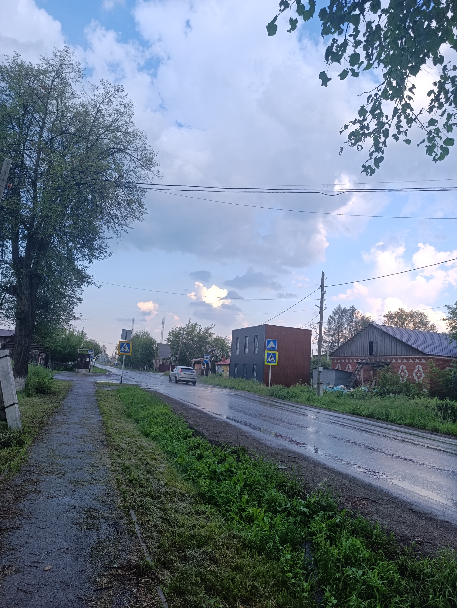 The image depicts a wet, asphalt road in a rural setting, possibly a village in Tugulym, Russia. The road appears to have recently been rained on, with visible puddles and reflections. On the left side of the road, there is a grey paved sidewalk with some grass and debris. To the right of the sidewalk is a grassy area with mowed grass. Beyond the sidewalk and grassy area, there are various structures. On the far left, a large tree with green leaves stands prominently. Further down the road on the left, a bus stop shelter is visible, with some white pillars.
Across the road, on the right side, there are several buildings. One is a modern, rectangular building with a dark facade and several windows. Next to it is a more traditional brick building with a gabled roof and decorative brickwork. The scene is bordered by green vegetation and trees.
A white car is driving down the wet road, heading away from the viewer. There are two pedestrian crossing signs with yellow backgrounds and black triangular symbols indicating a pedestrian. Overhead, power lines stretch across the frame, and the sky is filled with a mix of blue sky and white, fluffy clouds, suggesting it might be late afternoon or early evening, possibly after a rain shower. The overall mood is calm and a bit damp, reflecting the recent weather. No people are directly visible, but the presence of the car and bus stop implies ongoing activity.