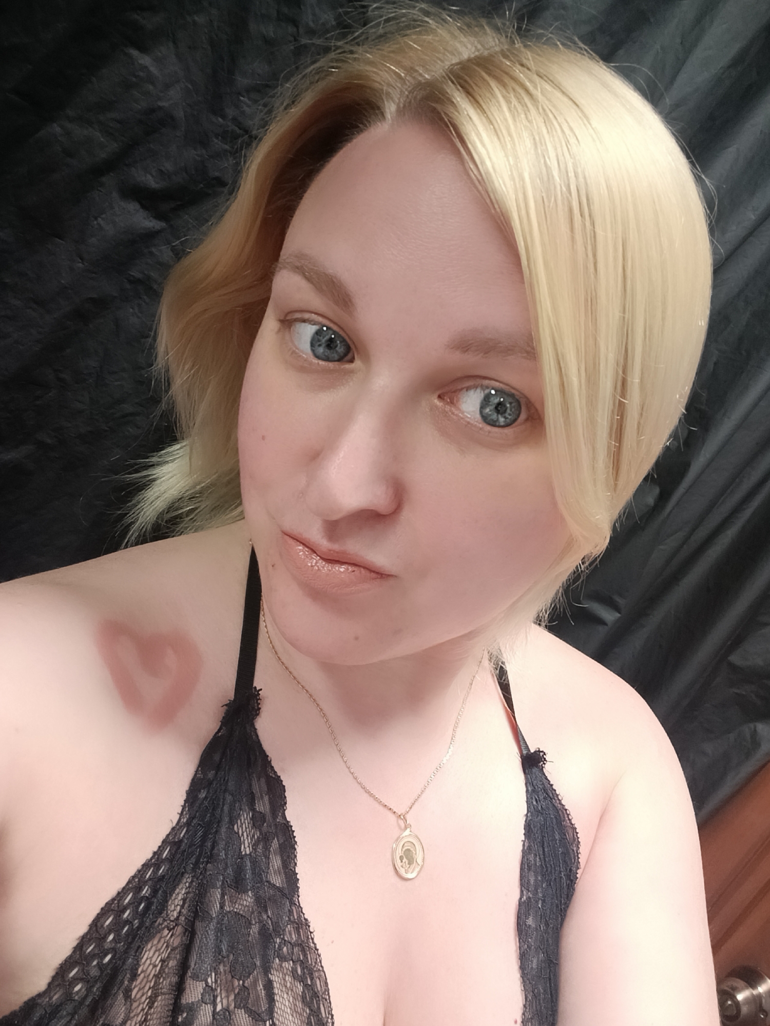 A close-up, chest-up portrait features a light-skinned individual with short, light blonde hair and clear blue eyes, set against a dark, crumpled fabric background. The person looks directly at the camera with a slightly pouting expression on their lips, which are coated in a glossy, light nude-colored lipstick. Their gaze appears playful or curious.
They are wearing a black lace bralette or camisole, with a thin strap visible over the shoulder. A delicate gold-colored chain necklace adorns their neck, featuring a small, oval pendant that appears to depict a religious figure. A distinct reddish, heart-shaped mark is visible on the left side of their chest, near the collarbone. The lighting suggests an indoor setting, likely with artificial light. There are no specific visual cues within the image to indicate the provided location of Chaikovsky, Russia. No text is visible in the frame.