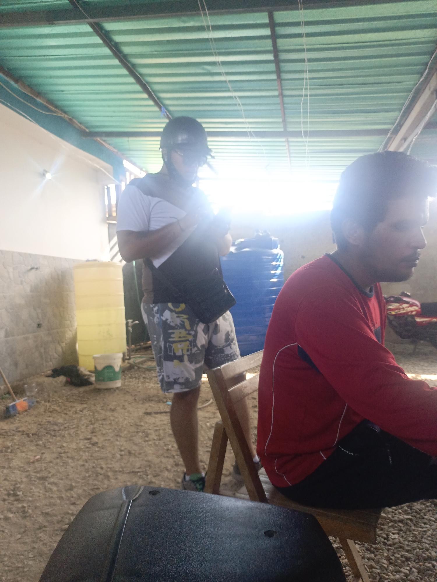 In Juan Griego, Venezuela, a man wearing a black helmet and sunglasses stands with his back to the camera, adjusting a black vest over a white shirt. He wears patterned shorts and athletic shoes. To his right, another man in a red long-sleeved shirt and black pants sits on a wooden chair, facing away from the camera. In the background, a large yellow water tank and a blue barrel are visible, along with a tiled wall. The ground is covered in gravel. The scene appears to be an outdoor or semi-outdoor area, possibly a workshop or storage space, with a corrugated metal roof overhead. A bright light source is positioned in the upper right of the frame, creating a lens flare.