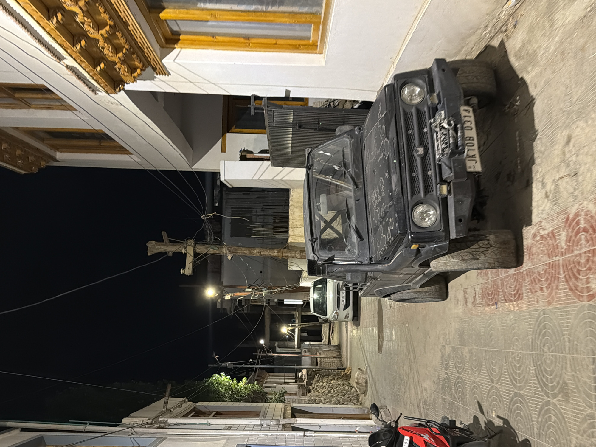 This is a night-time, eye-level, medium shot of a narrow street in Leh, India. In the foreground, a black, open-top SUV, likely a Suzuki Gypsy, is parked on the right side of the street, facing forward. Its license plate reads "JK08 0314". To the left, a red and black scooter is visible. The street is paved with patterned tiles.
In the background, a white car is parked further down the street, also facing forward. The street is lined with buildings on both sides. The building on the right has traditional Tibetan architectural elements, with ornate wooden carvings above the windows. The building on the left appears to be a guesthouse or shop with a stone wall base. There are several streetlights illuminating the scene, and power lines are visible overhead. A wooden utility pole with ceramic insulators stands prominently in the middle ground. A dog is seen lying on the ground in the shadow of the building on the left. The overall impression is a quiet, dimly lit urban environment.