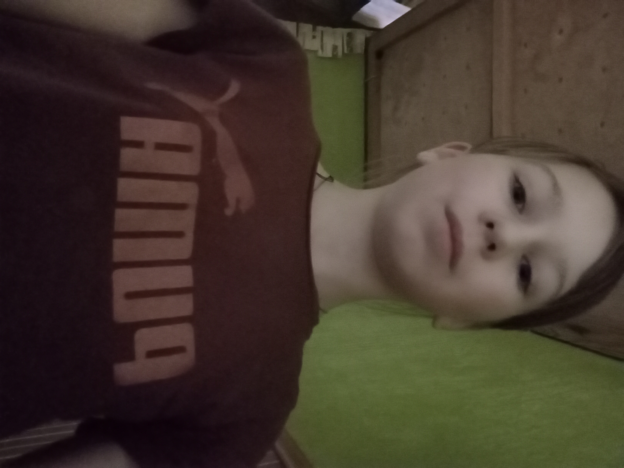 A child, viewed from a slightly low angle, wears a maroon Puma t-shirt with the Puma logo and name printed in light gray. The child is fair-skinned with brown hair and brown eyes. The background shows a light green surface and a wooden panel wall. Some objects hanging on a clothesline are partially visible in the background. The location in the city of Odesa, Ukraine is not discernible from the image.