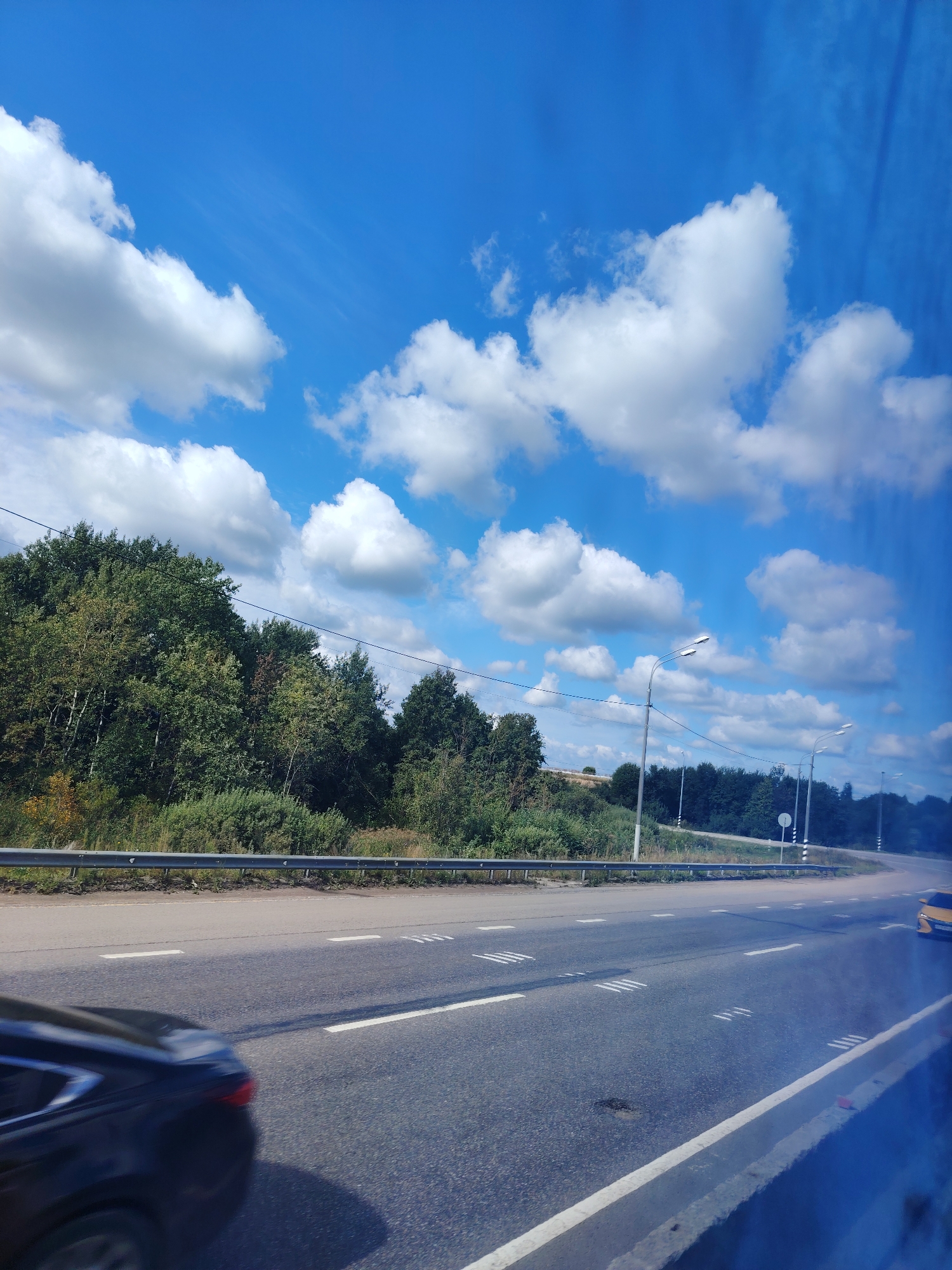 The image captures a roadside scene under a bright blue sky with scattered white cumulus clouds, suggestive of a sunny day. The viewpoint appears to be from inside a moving vehicle, looking out at a highway. A black car is visible in the foreground, moving away from the camera. The highway itself has multiple lanes marked with white lines, and some rumble strips are present. Along the side of the road, there is a metal guardrail, behind which is a dense line of green trees and foliage. Further in the distance, more trees and vegetation are visible, along with several streetlights. The sky dominates the upper portion of the image. The location is identified as the city of Крот Снт, Russia.