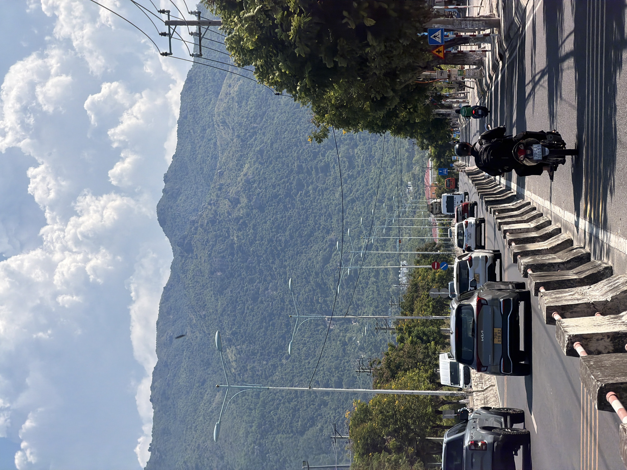 The image depicts a street scene in Nam Nha Trang Ward, Vietnam, on a sunny day with a blue sky filled with white clouds. A multi-lane road is visible, with concrete barriers separating the lanes. Several vehicles are present, including cars and motorcycles. On the left side of the road, a grey pickup truck is driving, followed by a grey SUV with a license plate ending in "078.92". Further ahead, a white van and other cars are visible. On the right side of the road, a person is riding a motorcycle, wearing a black jacket and helmet, and a backpack. Another motorcycle is further down the road. Trees line the right side of the street, and utility poles with wires are also present. In the background, a large, green, tree-covered mountain dominates the landscape. Buildings and infrastructure are subtly visible along the road.