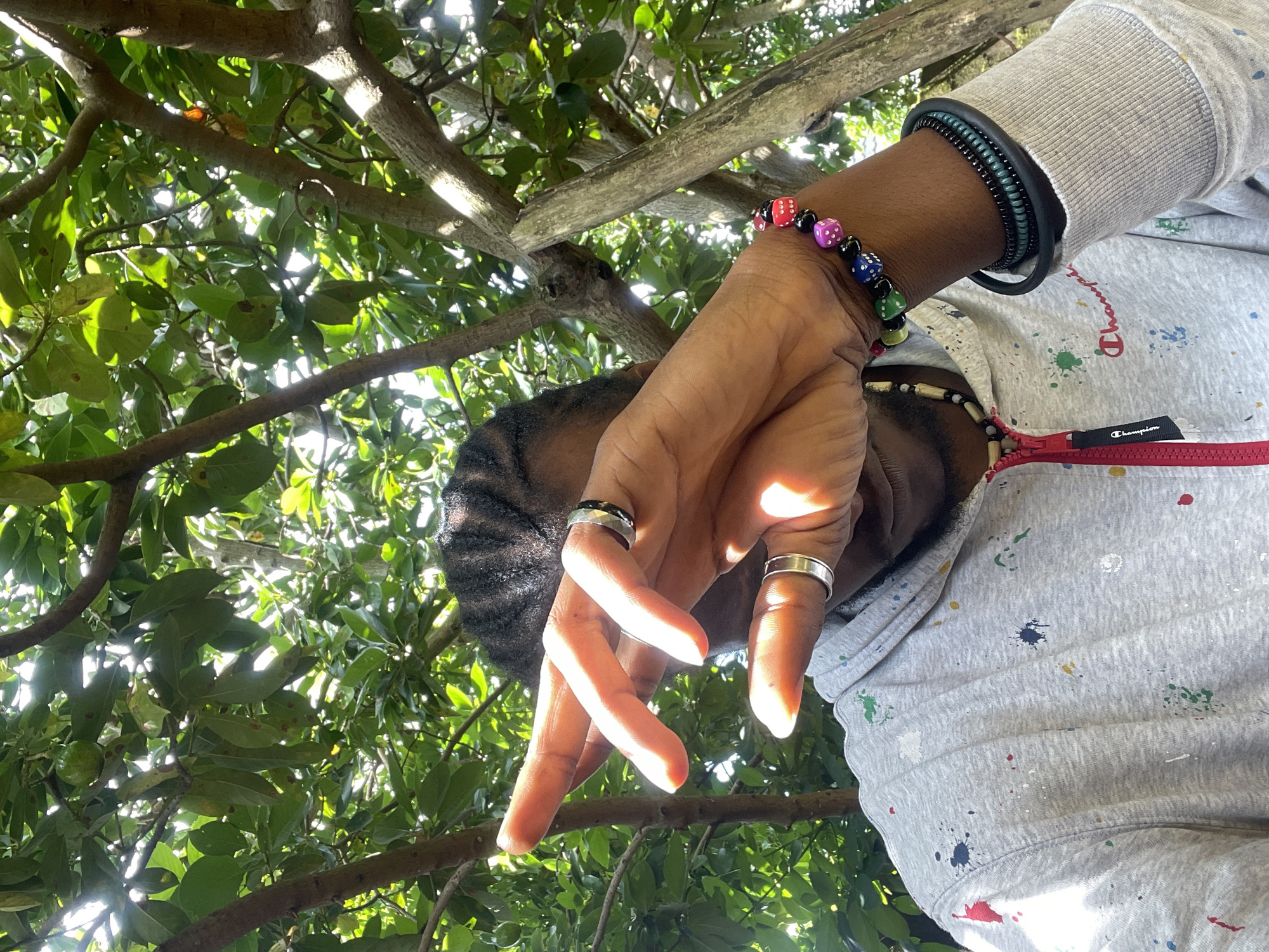 A person is captured in a medium shot, standing outdoors under a canopy of green foliage, likely in a garden or park. The person's face is partially obscured by their hand, which is raised to cover their eyes, but their left arm is visible, adorned with a colorful beaded bracelet featuring dice and other small charms. They are wearing a gray hooded sweatshirt with a red zipper, decorated with various paint splatters in red, blue, green, yellow, and white. Two silver rings are visible on their fingers. The background consists of tree branches and leaves, with sunlight filtering through, creating dappled light and shadows. The atmosphere suggests a casual, outdoor setting. The question of this scene's location within the city of Jos, Nigeria cannot be determined from the visual information provided.