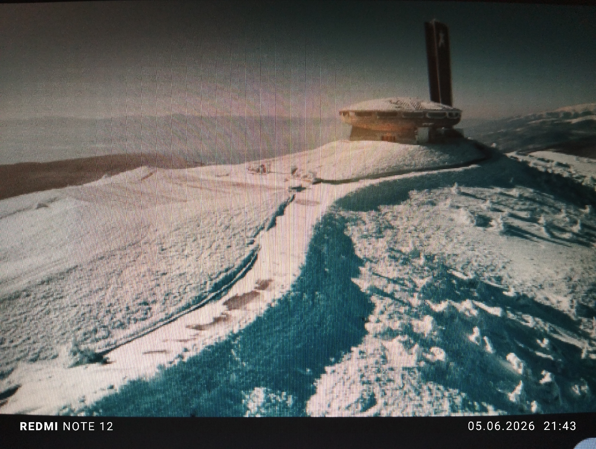 The image depicts the Buzludzha Monument, located on Buzludzha Peak in the Central Balkan Mountains, Bulgaria. This location is approximately 200 kilometers northwest of Burgas, Bulgaria, contradicting the specified city in the prompt.
The scene presents an outdoor, high-altitude landscape dominated by the snow-covered Buzludzha Monument under what appears to be a late evening or early morning sky. The monument, a concrete, saucer-shaped structure with a prominent tall pylon on its left side, is situated on a snow-covered rise. Its exterior surfaces exhibit a dark, possibly reddish-brown tone, contrasting with the white snow that partially covers its roof and the surrounding ground.
The foreground and midground consist of undulating terrain blanketed in white snow. A distinct, winding path or set of tracks is visible in the snow, leading from the lower left towards the base of the monument. Portions of the snow exhibit bluish tints, likely due to shadows or lighting conditions. The sky, occupying the upper portion of the frame, is overcast or hazy, with a gradient from a lighter gray near the horizon to a darker gray-blue overhead. Faint outlines of distant snow-covered mountains or hills are discernible on the horizon to the right of the monument.
No individuals, animals, or active vehicles are present within the frame. The image itself displays visible vertical and horizontal lines, characteristic of a photograph captured from a digital display screen. A black bar at the bottom contains white text: "REDMI NOTE 12" on the left and "05.06.2026 21:43" on the right, indicating the capture device and timestamp.