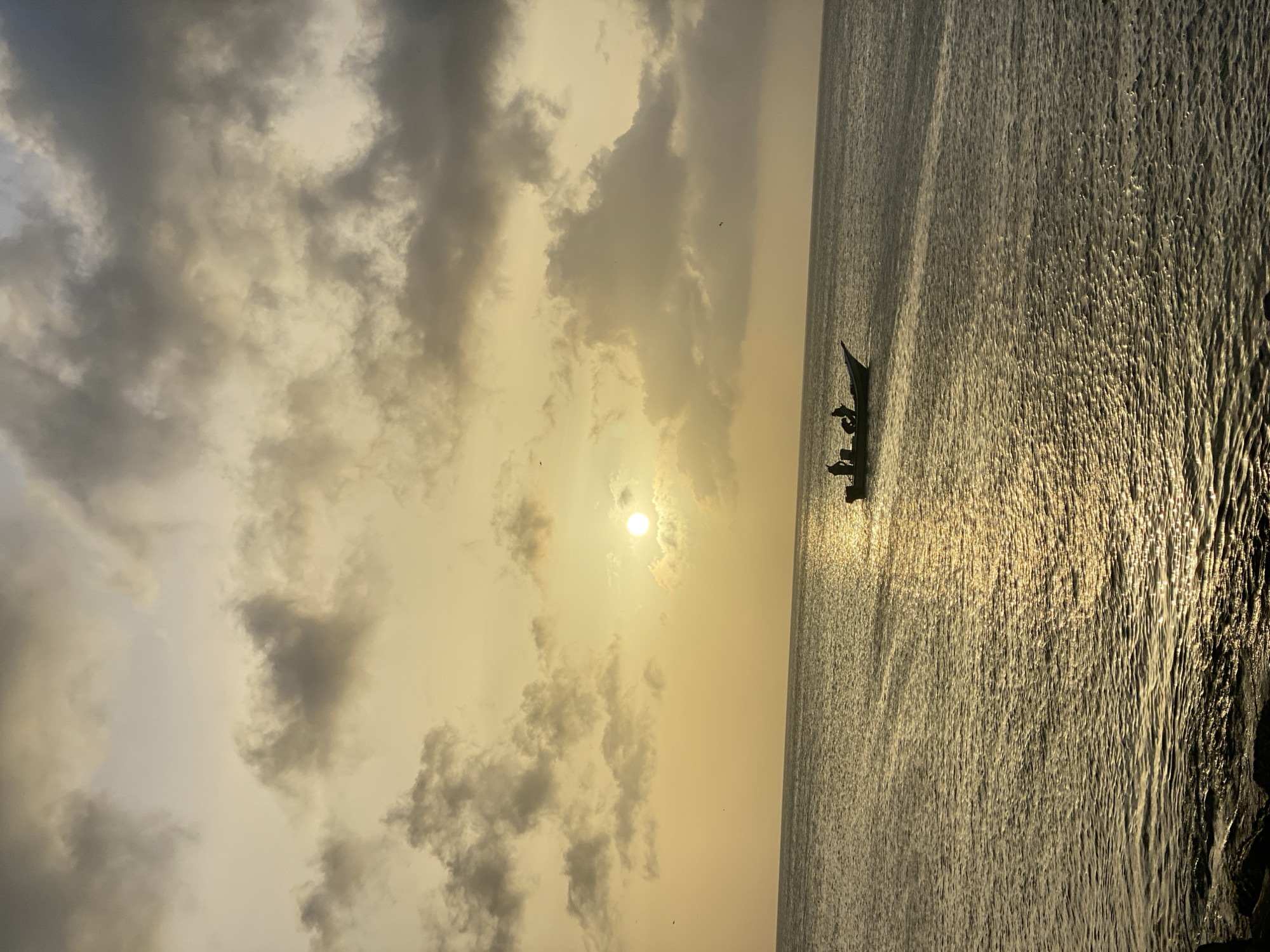 The image shows a sunset scene in Juan Griego, Venezuela. The sun is partially obscured by clouds. A small boat with several people is visible on the water. The sea is calm with reflections of sunlight. The sky is filled with clouds.