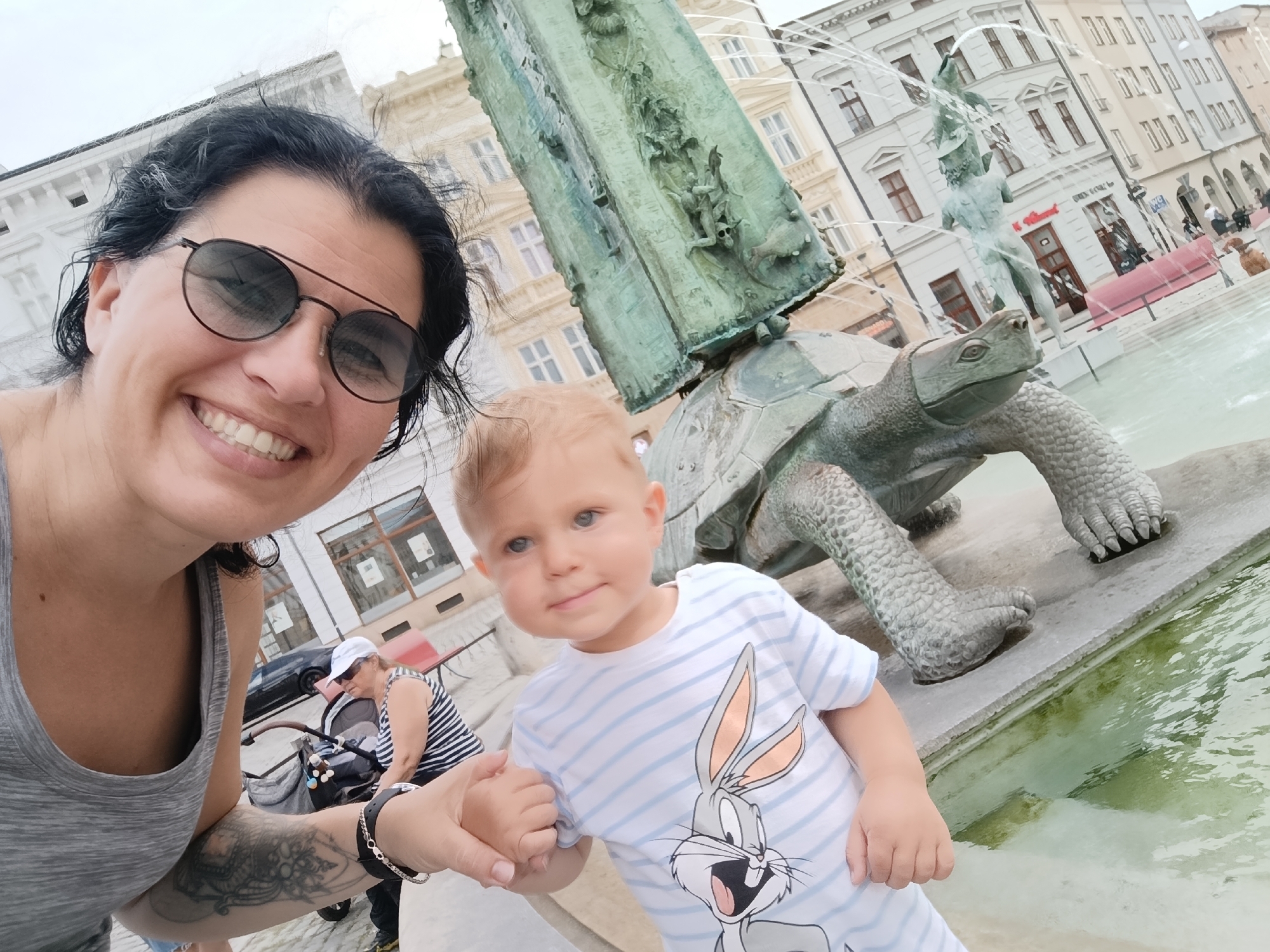 A woman and a young child are taking a selfie in front of a fountain in Olomouc, Czechia. The woman, on the left, is wearing sunglasses and a grey tank top. She has dark hair and is smiling. The child, on the right, is wearing a striped t-shirt with a Bugs Bunny graphic. They are both in focus and positioned in the foreground of the image. Behind them, a large, ornate fountain featuring a turtle sculpture is visible, with water spraying from it. The background shows a street with historical buildings, typical of Olomouc's city center. An older woman and a stroller are also visible in the background, to the left of the fountain.