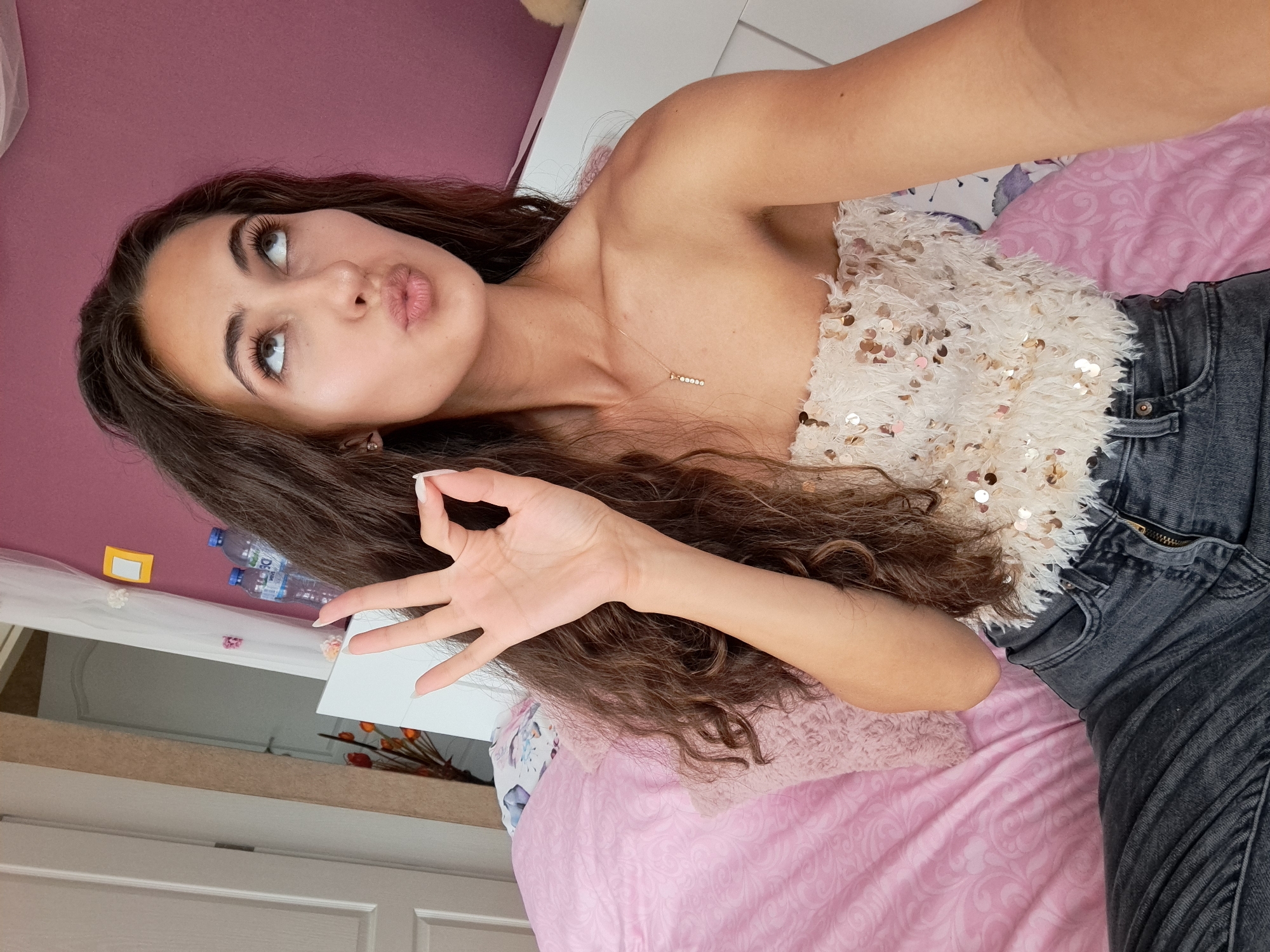 A young woman with long, dark brown, curly hair is seated indoors, likely in a bedroom, capturing a selfie. She performs an "OK" hand gesture with her left hand, with long, light-colored nails. Her lips are pursed in a "kissy" expression, and her eyes are rolled upwards and to the right. She is wearing a strapless, light-colored top adorned with iridescent sequins and a feathery texture, paired with dark grey denim jeans. A delicate gold-toned necklace with a vertical, multi-stone pendant is visible on her chest. In the background, to the left, is a white door next to a beige-textured wall, and a partial reflection of another door and a red-orange floral arrangement in a mirror. To the right, a dark pink wall is visible, along with a white headboard or piece of furniture on which two plastic water bottles are placed. A sheer white curtain with small floral decorations hangs on the right, and a small yellow rectangular object is affixed to the pink wall. A pink patterned bedspread and a fuzzy pink pillow are visible on the bed. The location is the city of Unknown, Unknown.
